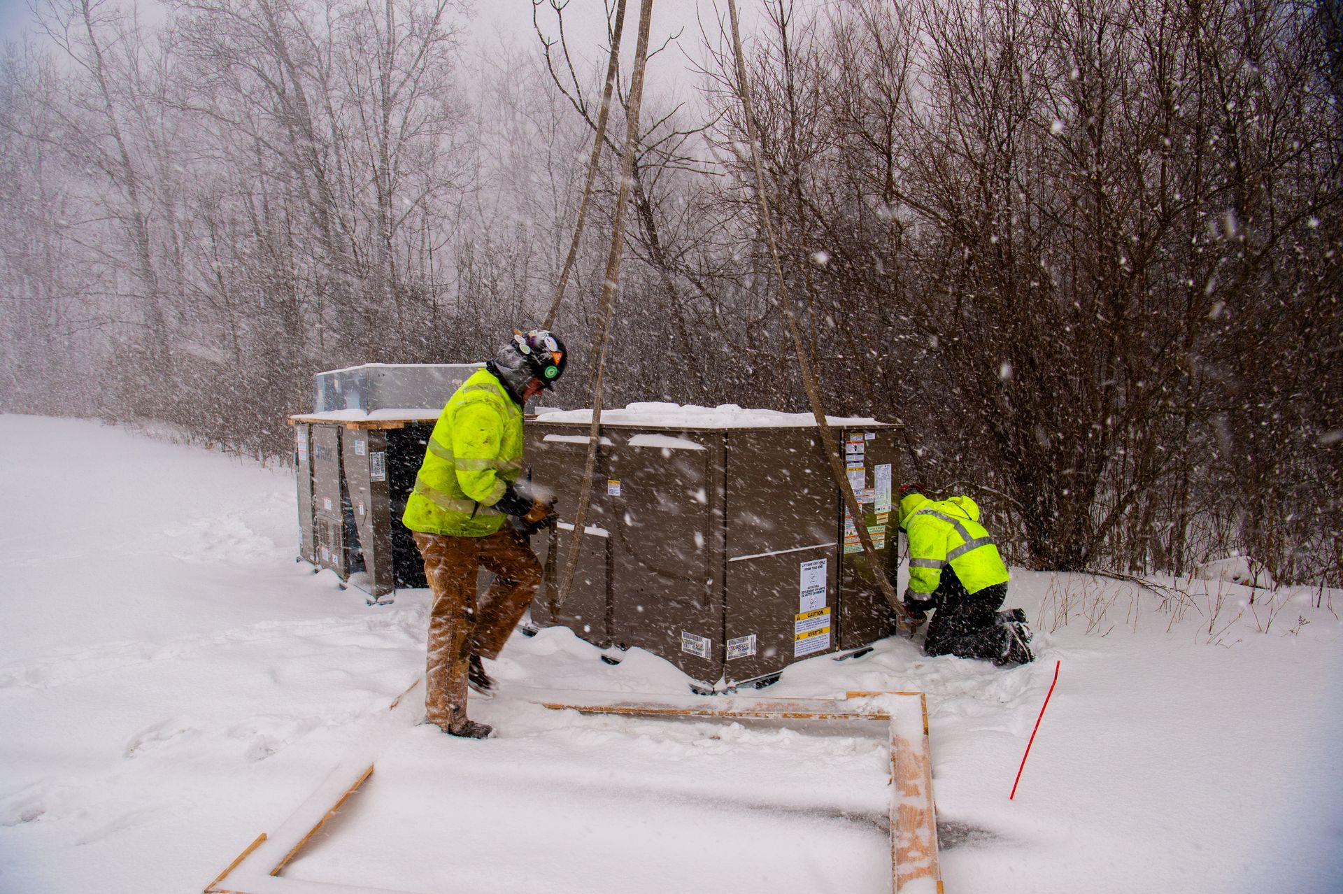 Two workers in winter gear building something outdoors; snow falling.