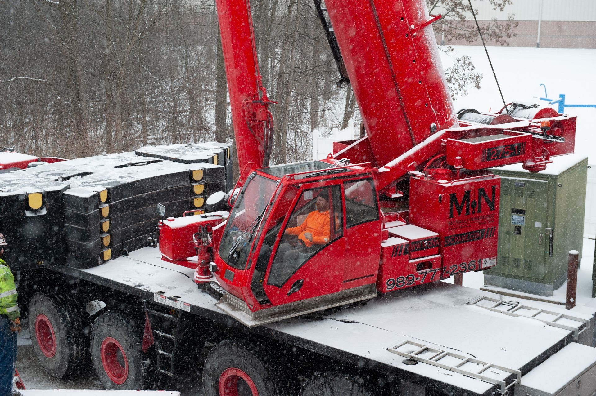 Red crane on a snowy day. Crane operator in cab. Black counterweights and a green utility box visible.