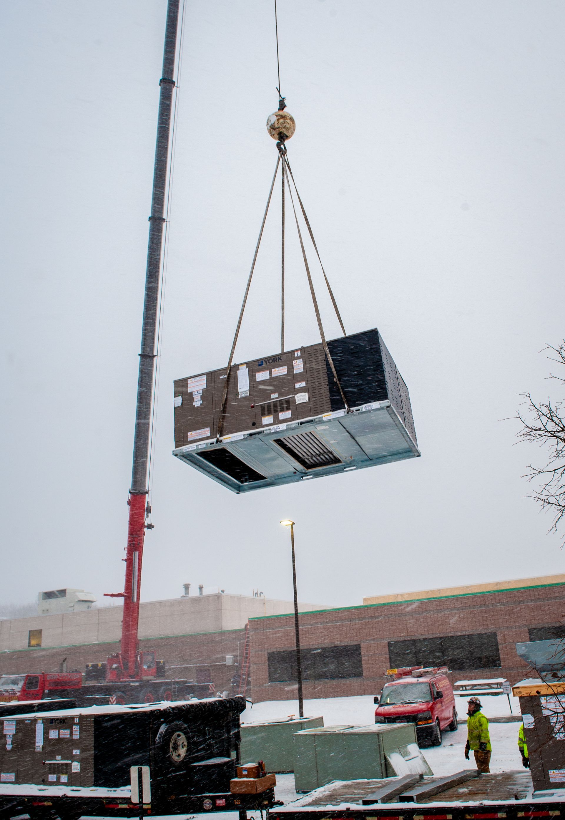 Crane lifting an HVAC unit on a snowy day near a building.