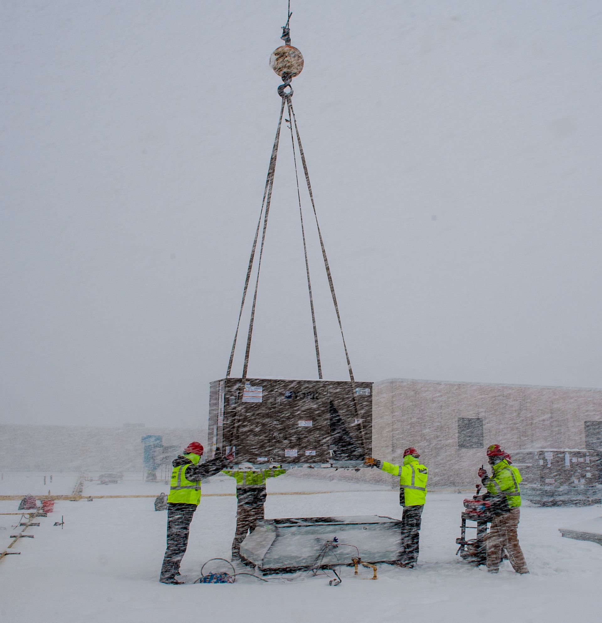 Workers in yellow vests lift a large crate with a crane in a snowy outdoor setting.