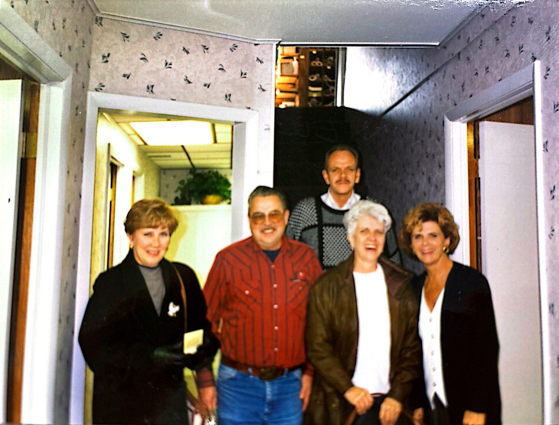 Five people in a hallway; three women and two men. Floral wallpaper, doorway with plants, and a light-filled room.