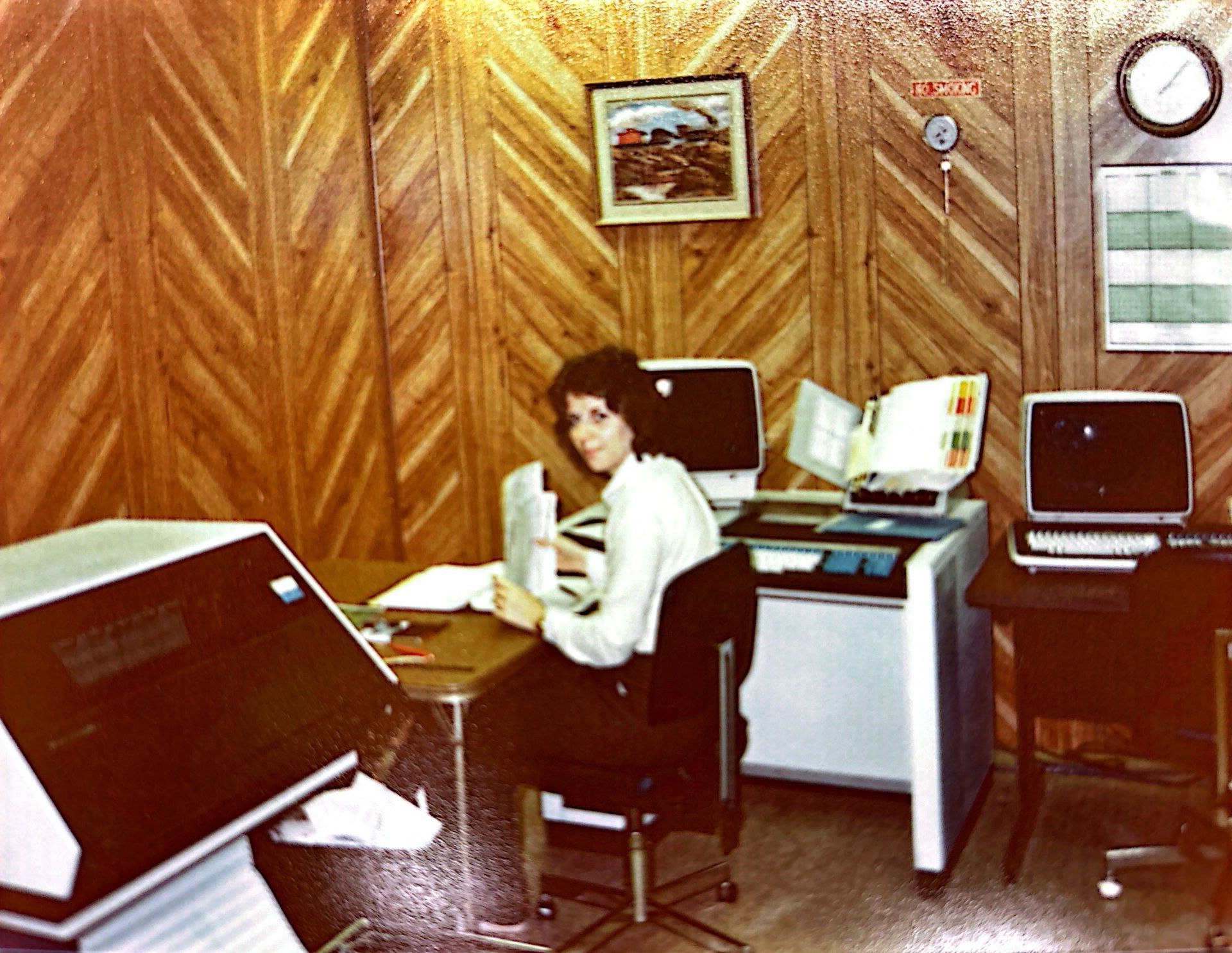 Woman seated at desk with vintage computer equipment in wood-paneled office.