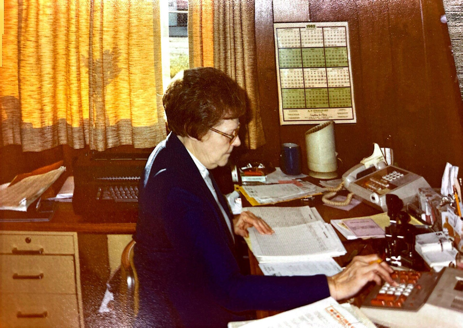 Woman at desk, working on documents and calculator; office setting with curtains and calendar.