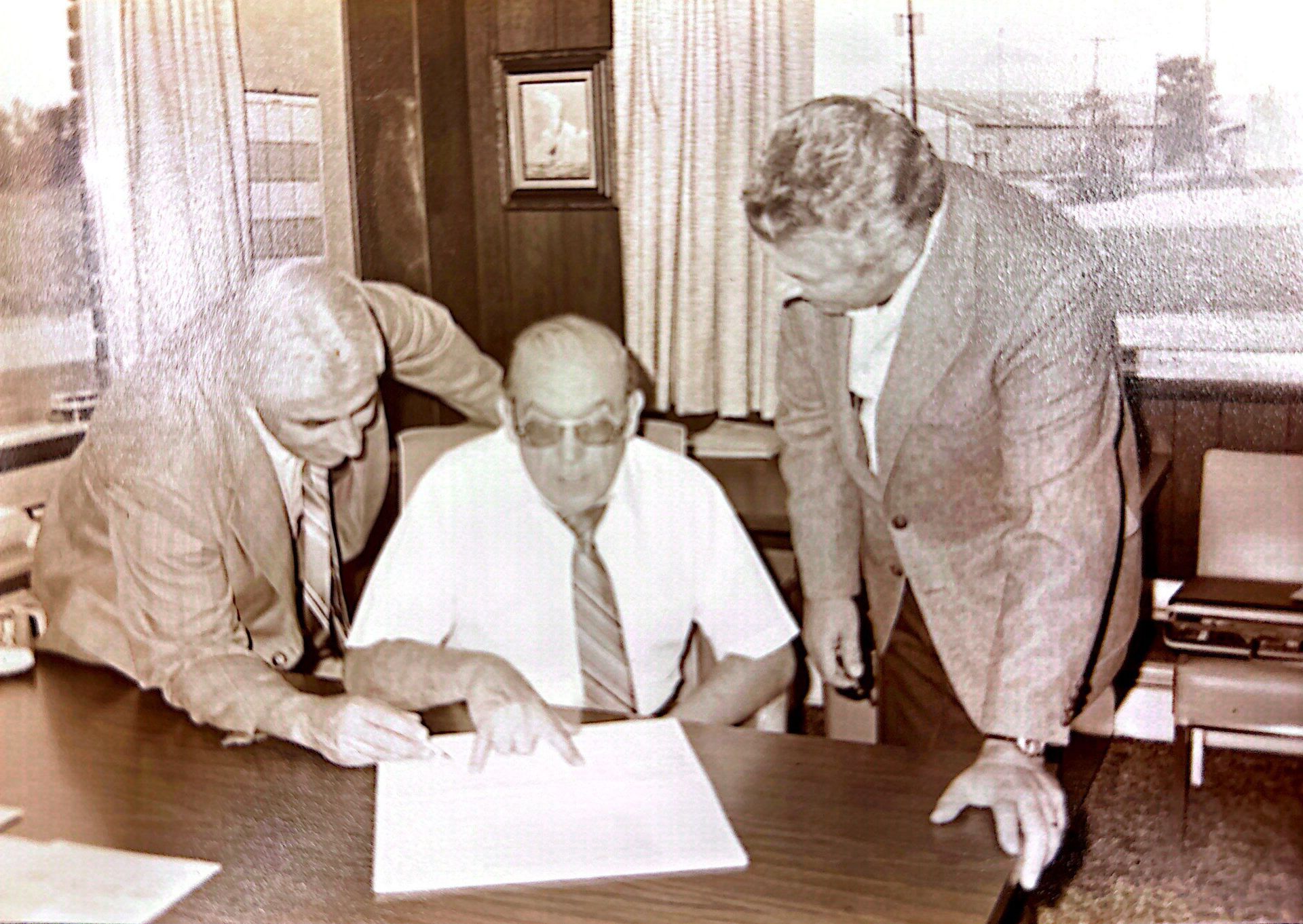 Three men at a desk, looking at a document. Two men point, the third sits, all wearing suits.