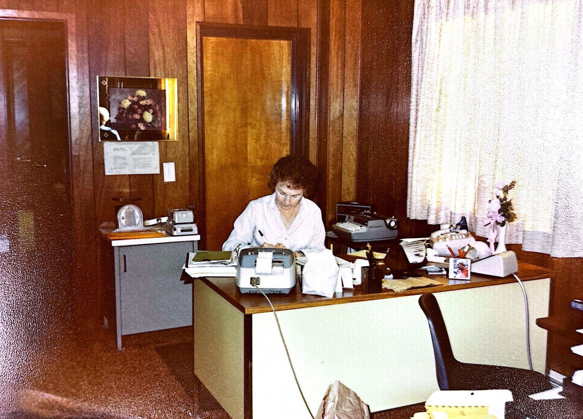 Woman in lab coat sits at desk in office with typewriter and other office supplies.