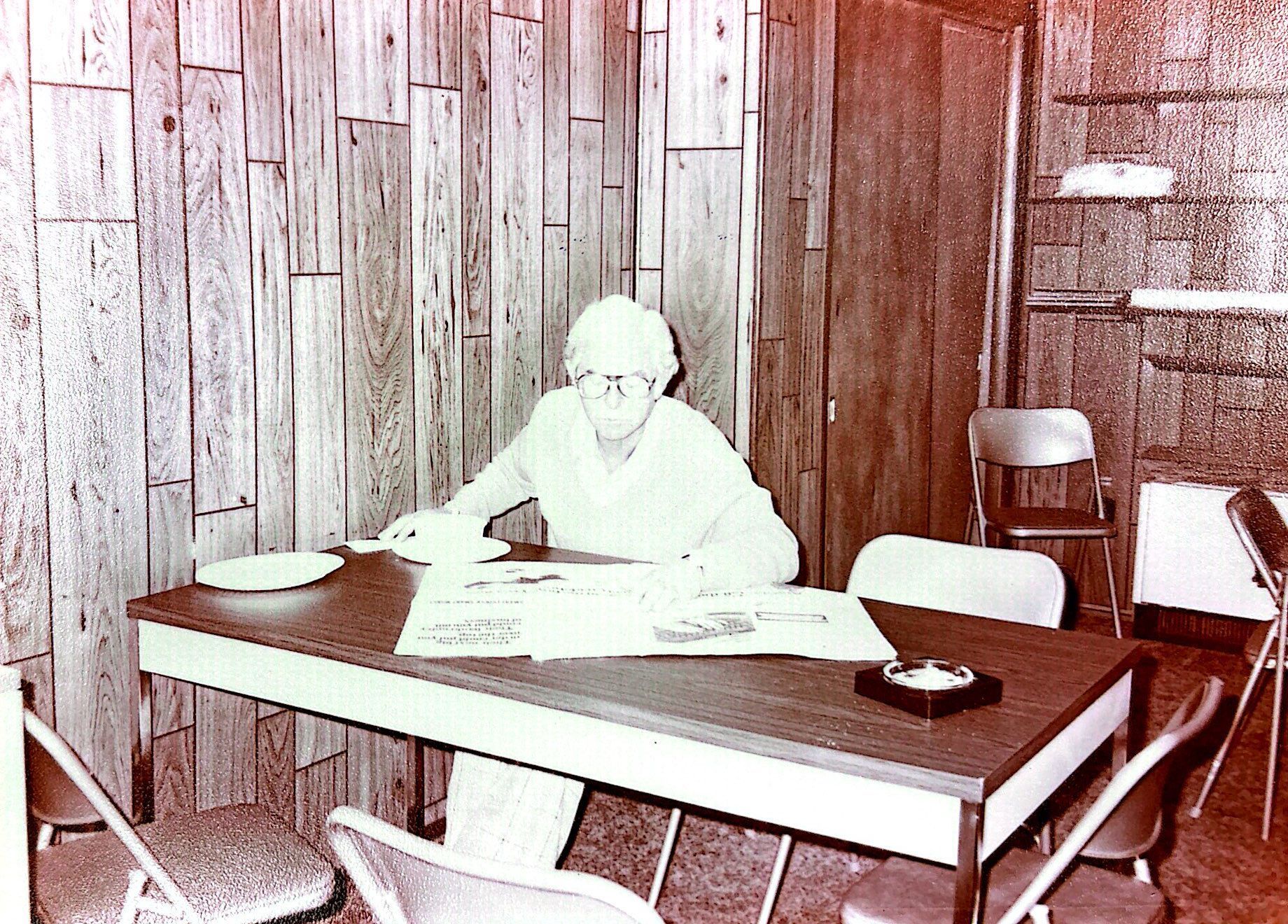 Person at desk in a wood-paneled room.  Several chairs surround the desk with papers and an ashtray.