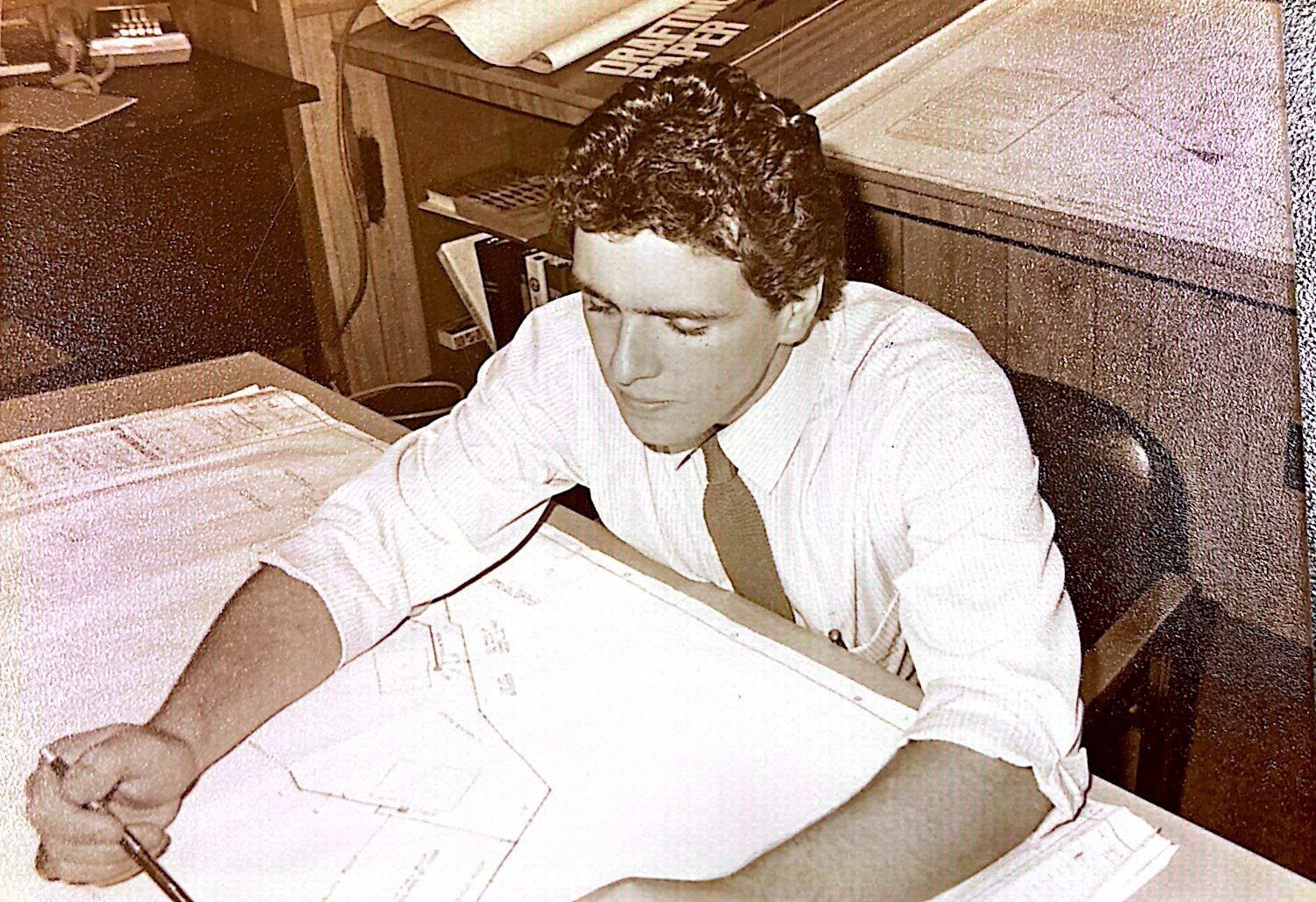 Man at a drafting table, focused, drawing. Wearing a light shirt and tie, in an office setting.