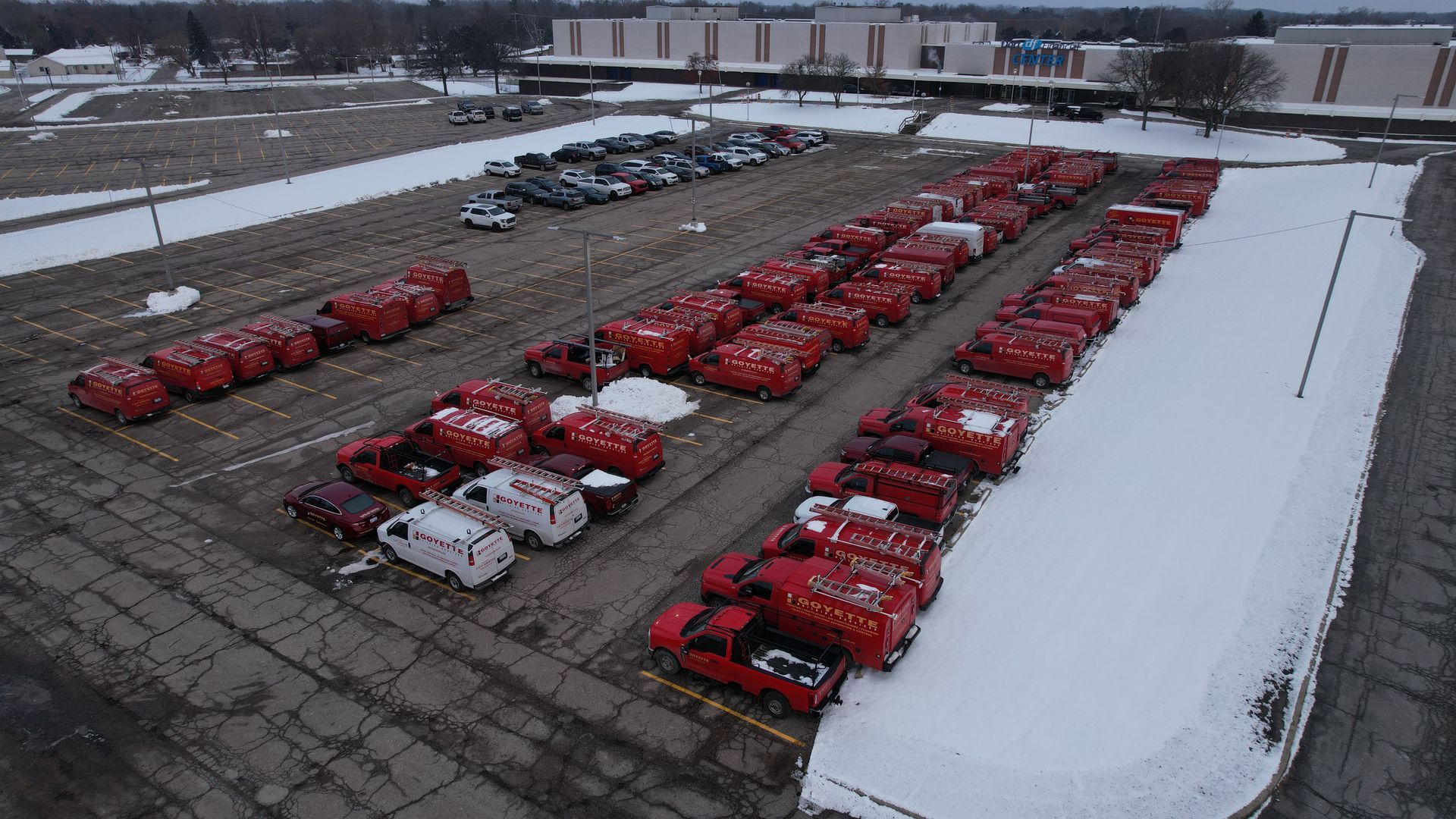 Aerial view of a snowy parking lot filled with red delivery trucks and vans parked in rows.
