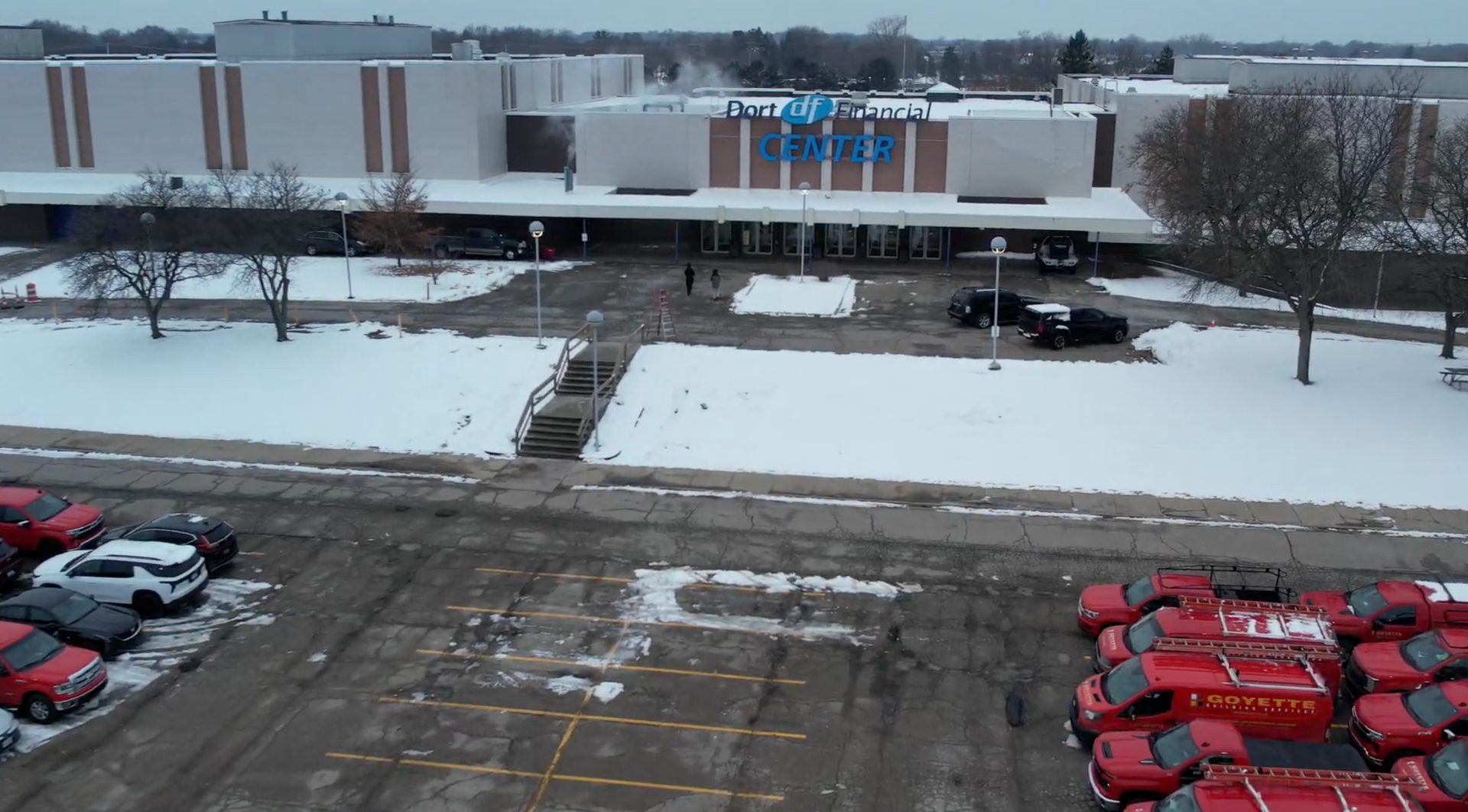 Exterior of a shopping mall entrance, snowy winter scene, red vehicles parked in the foreground.