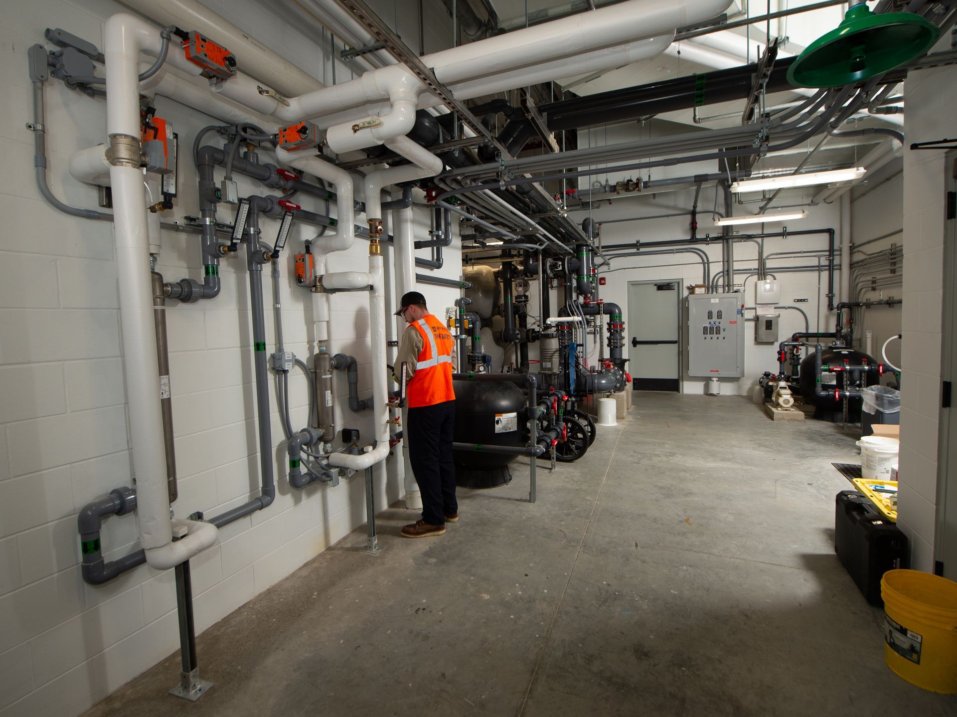 Person in orange safety vest in a utility room with pipes, machinery, and industrial equipment