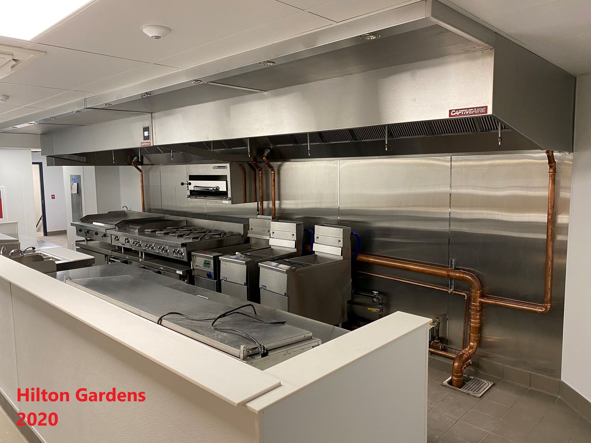 Commercial kitchen with stainless steel appliances and ventilation hood. Copper pipes visible. 