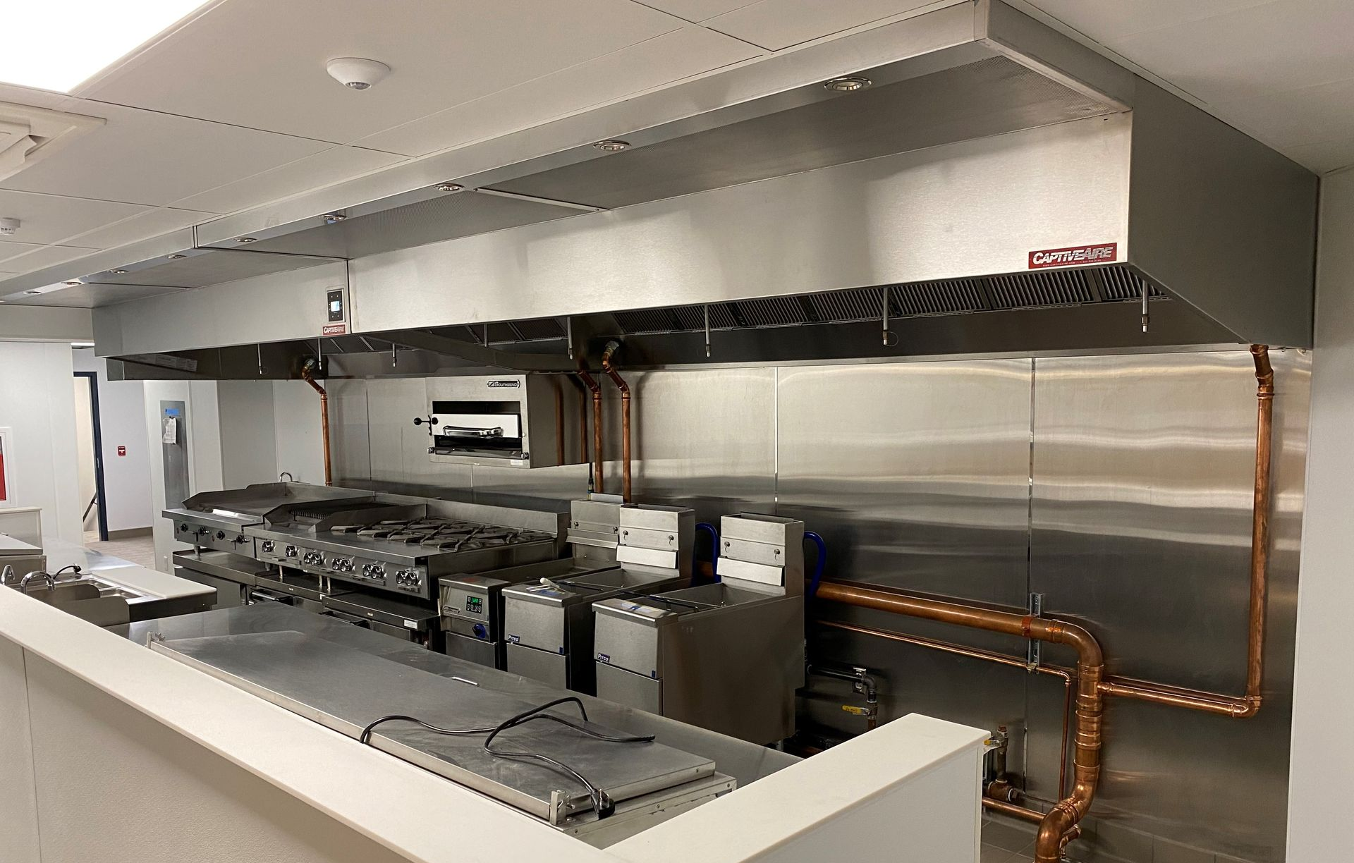 Commercial kitchen with stainless steel appliances, ventilation hood, and copper pipes.