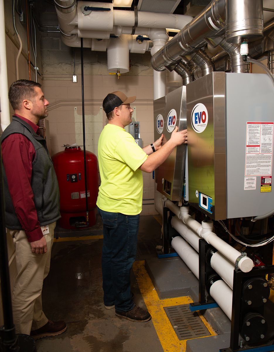 Two people in a mechanical room, one adjusting equipment, one observing. Pipes and a red tank are visible.