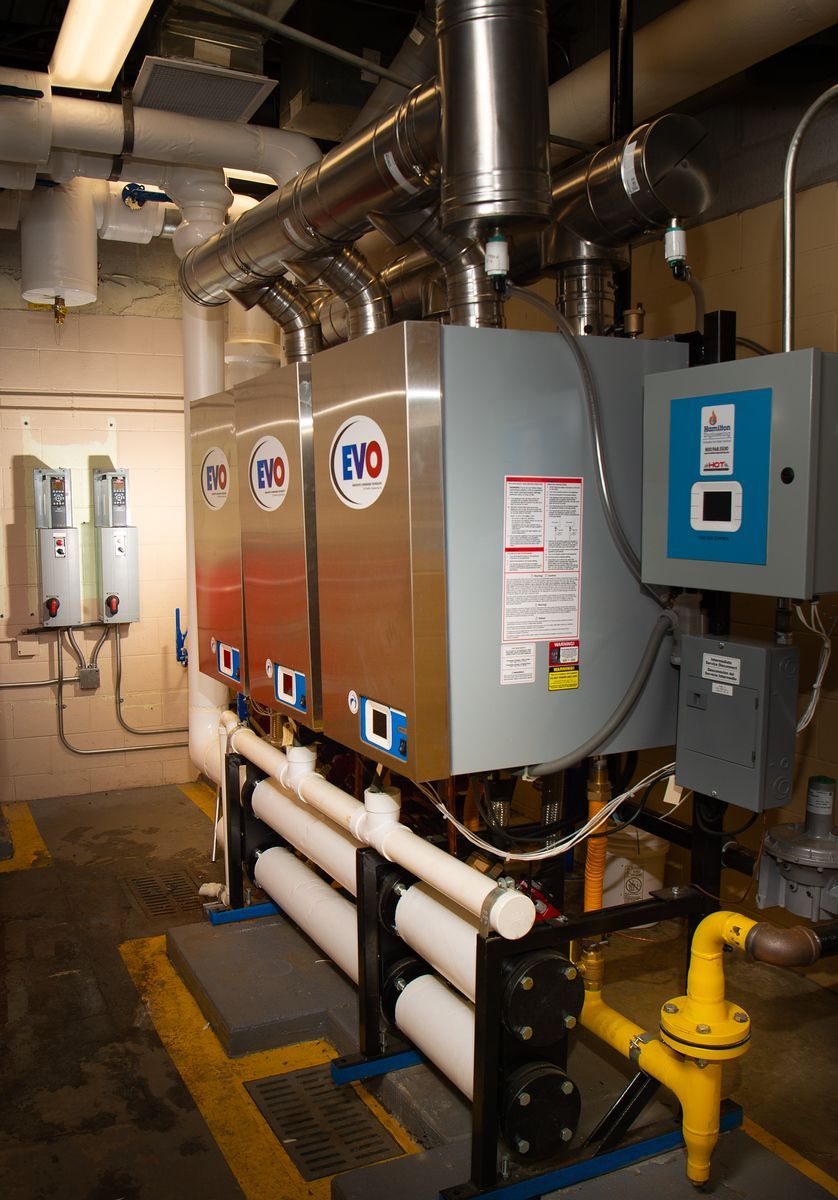 Three large, silver EVO water heaters in a utility room. Yellow gas pipe and white PVC pipes are visible.