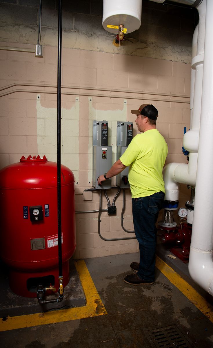 Man in safety vest operates control panel near red tank and pipes. Yellow caution lines mark the floor.