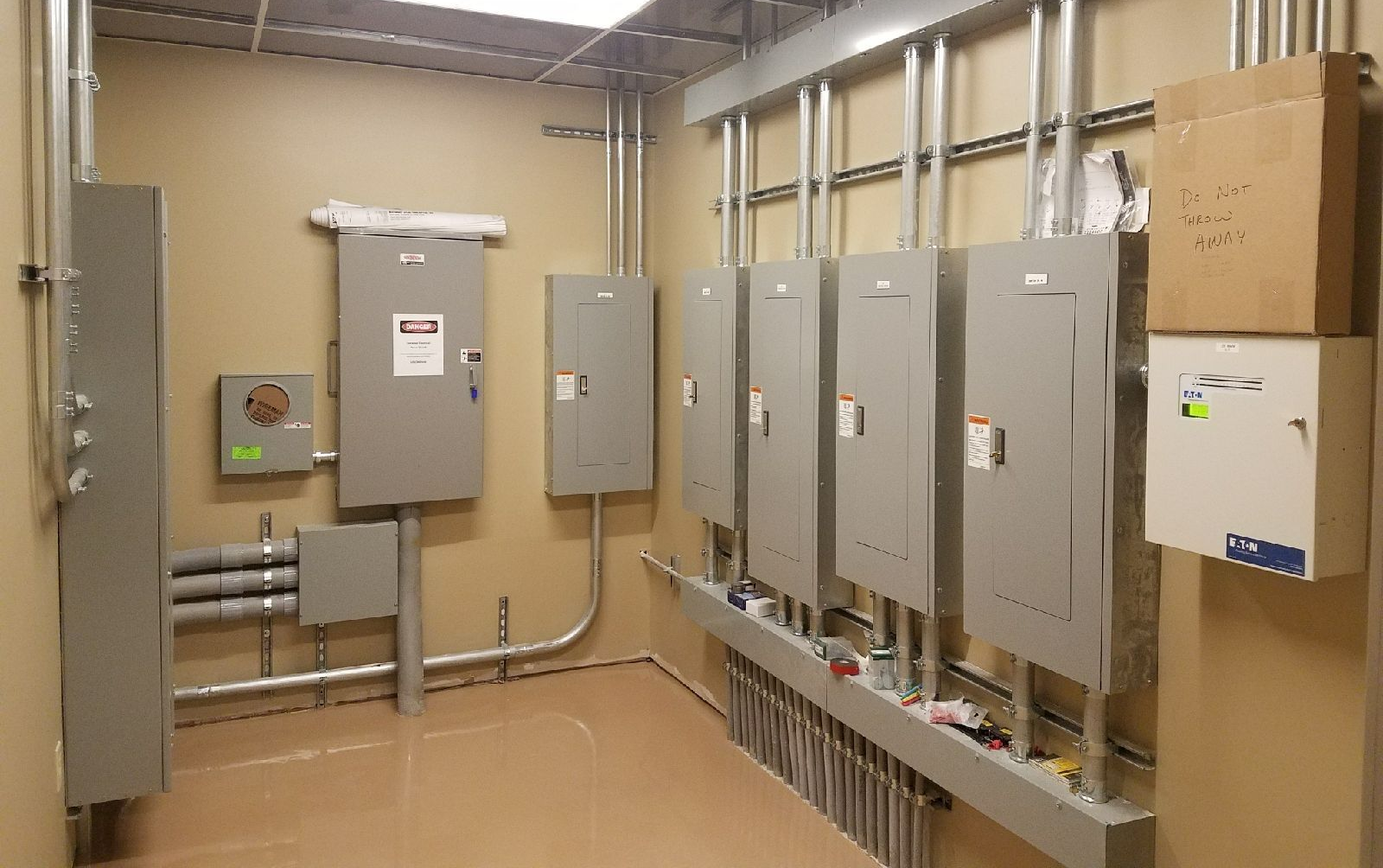 Electrical room with gray panels, conduits, and equipment against beige walls.