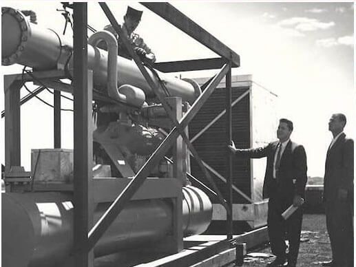Men inspecting machinery outdoors. One man in suit, two men working. Metal structure, sunny day.