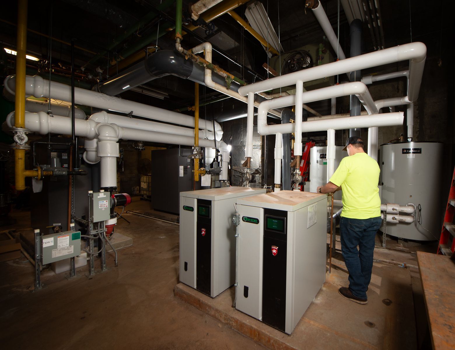 Man in a yellow shirt examines two industrial boilers in a mechanical room, surrounded by pipes.