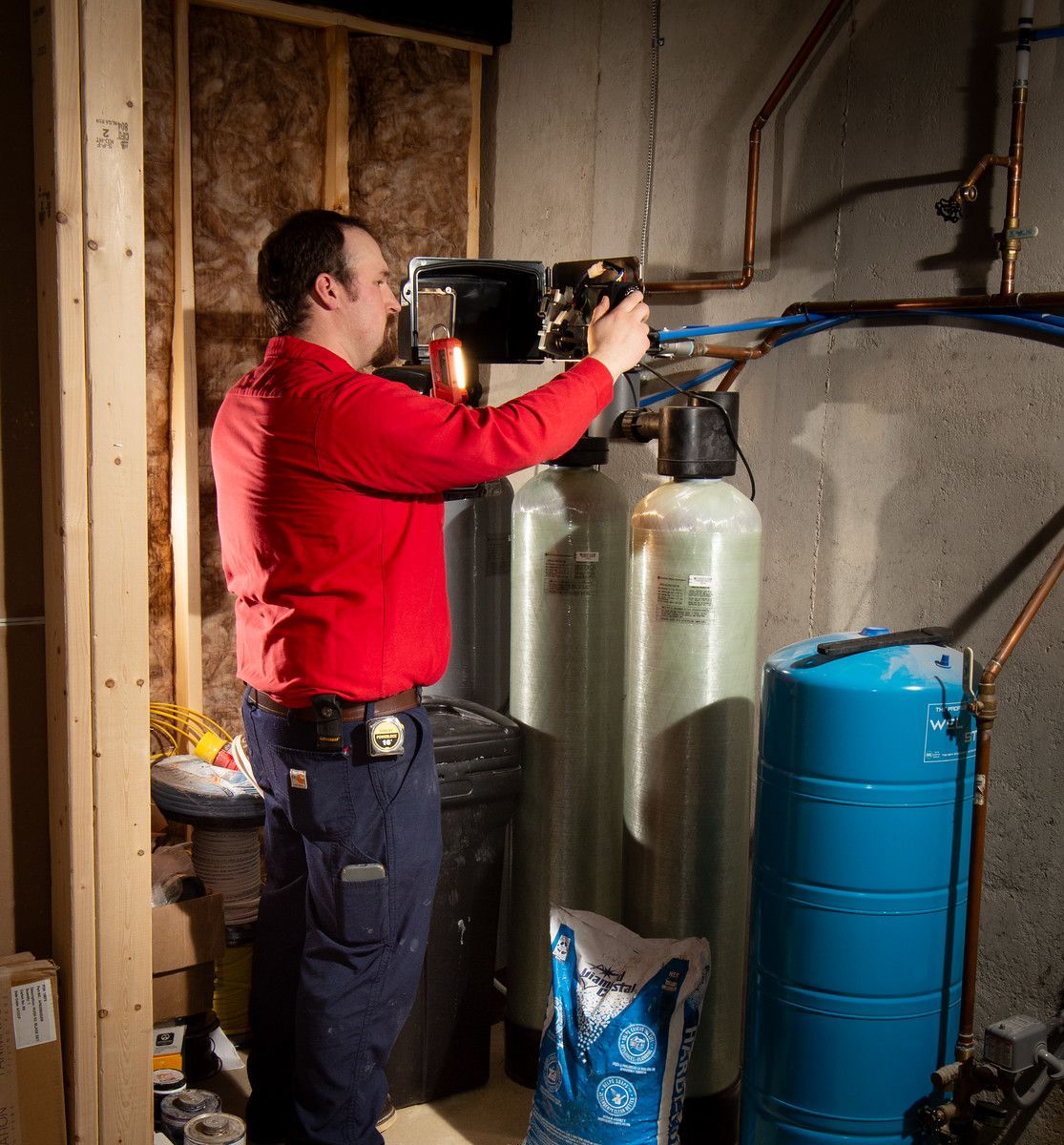 Plumber in red shirt installing water softener system in a basement.