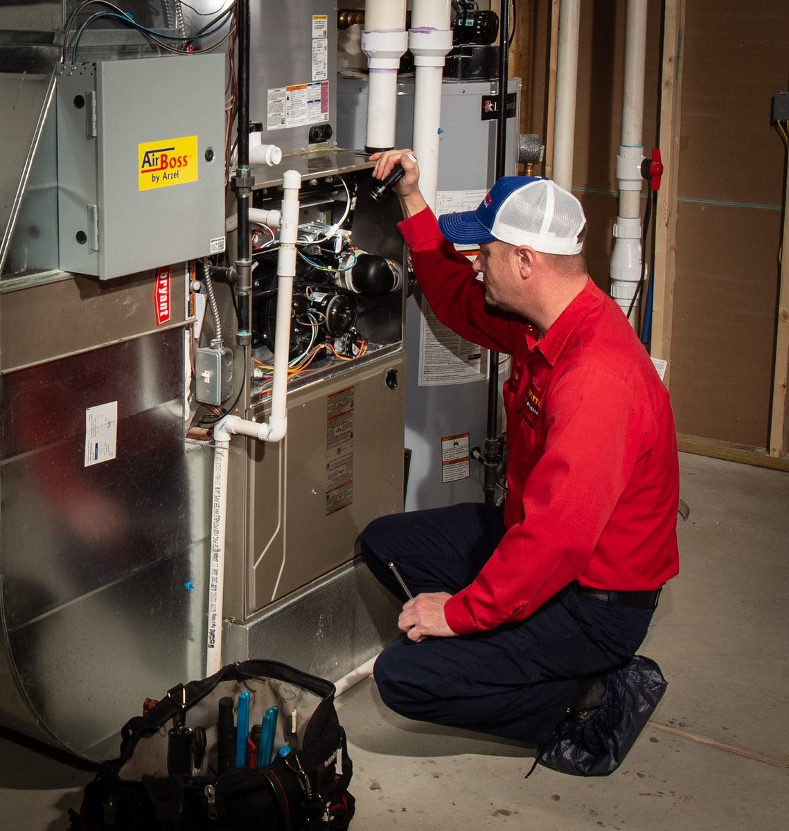 HVAC technician inspecting a furnace in a utility room. He is kneeling, wearing a red shirt and blue hat.