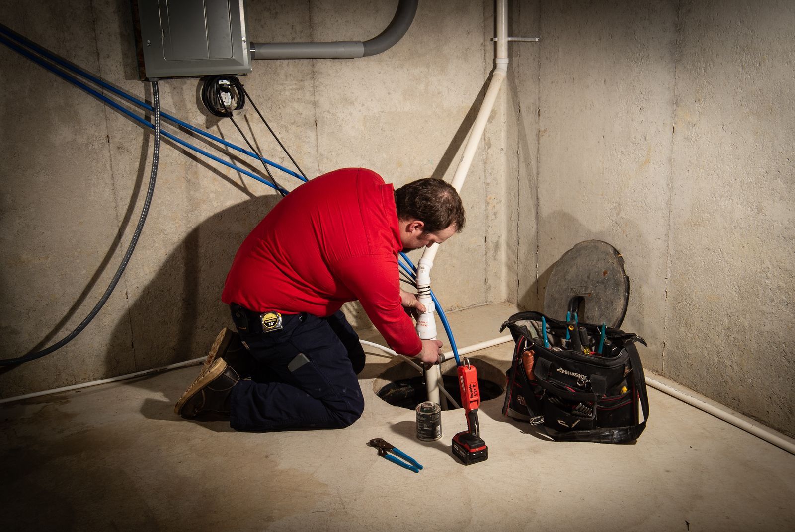 Plumber in red shirt kneels, working on pipes in a concrete basement. Tools and bag are nearby.