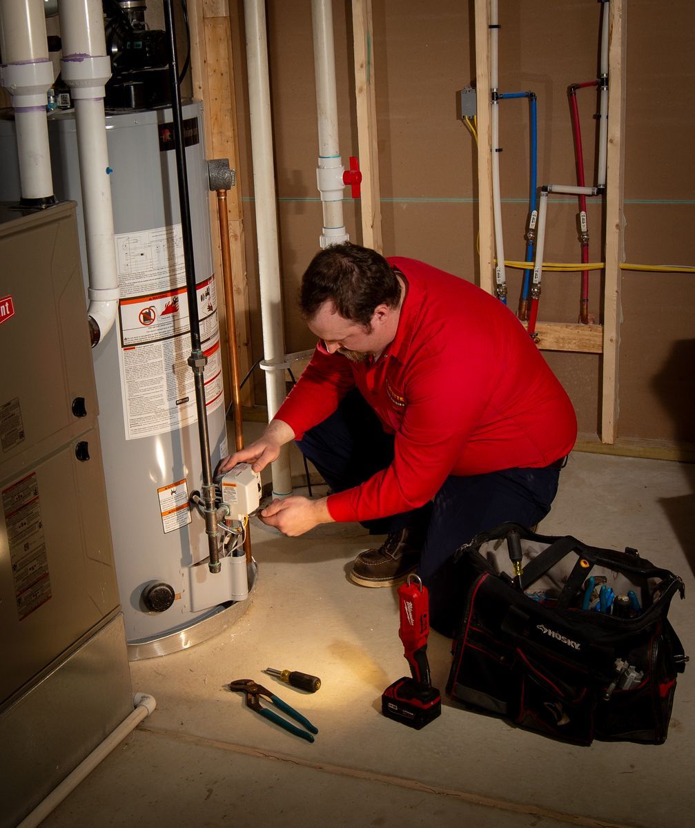 Plumber in red shirt working on a water heater in a basement. Tools in a bag and on floor.