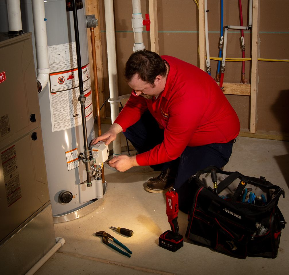 Plumber in red shirt examines water heater in a basement, tools nearby.