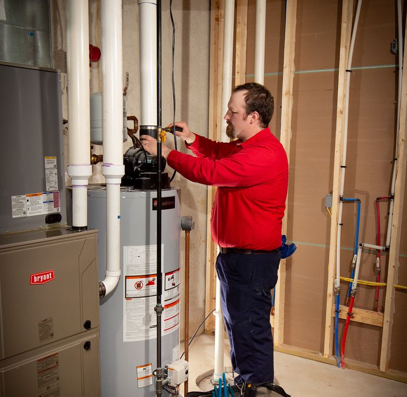 A plumber in a red shirt working on a water heater in a utility room.