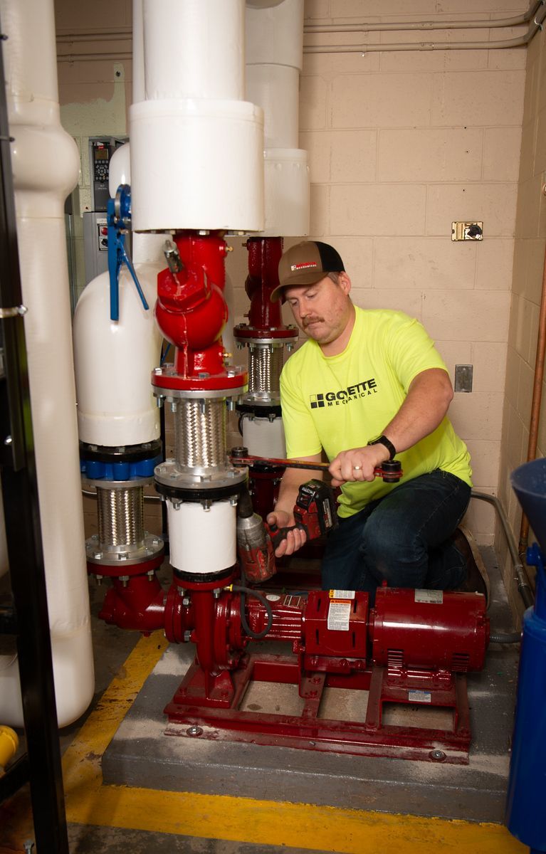 Man in a yellow shirt works on a red water pump in a mechanical room with pipes.