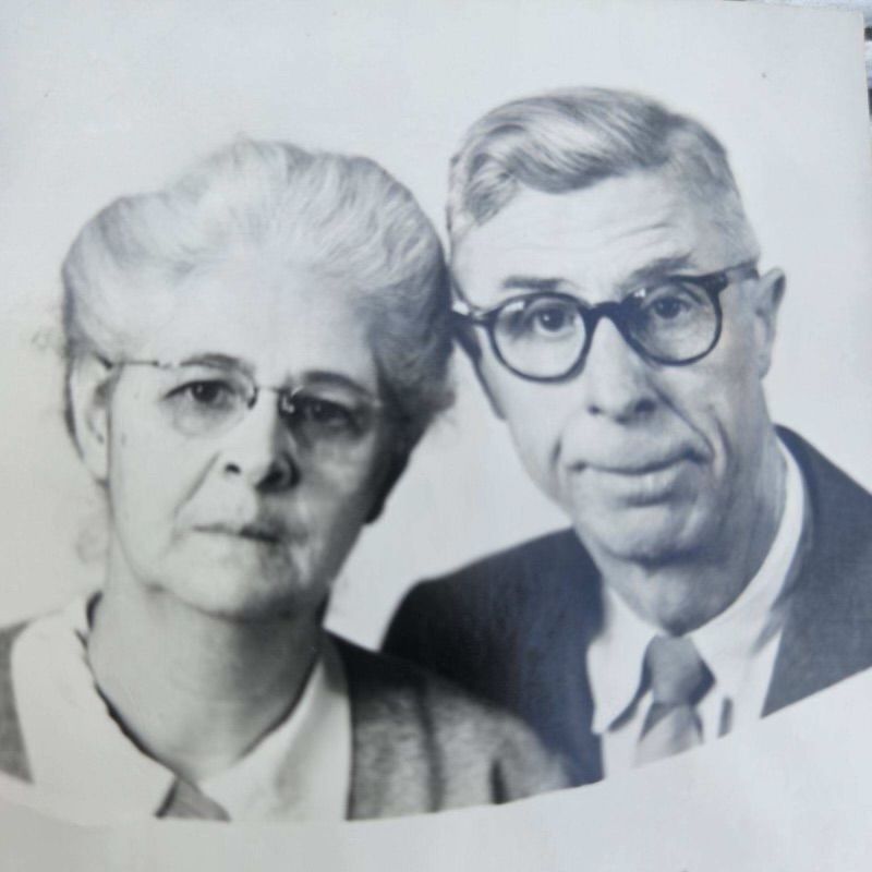 Portrait of an elderly couple, both wearing glasses. Woman in cardigan, man in suit. Black and white.