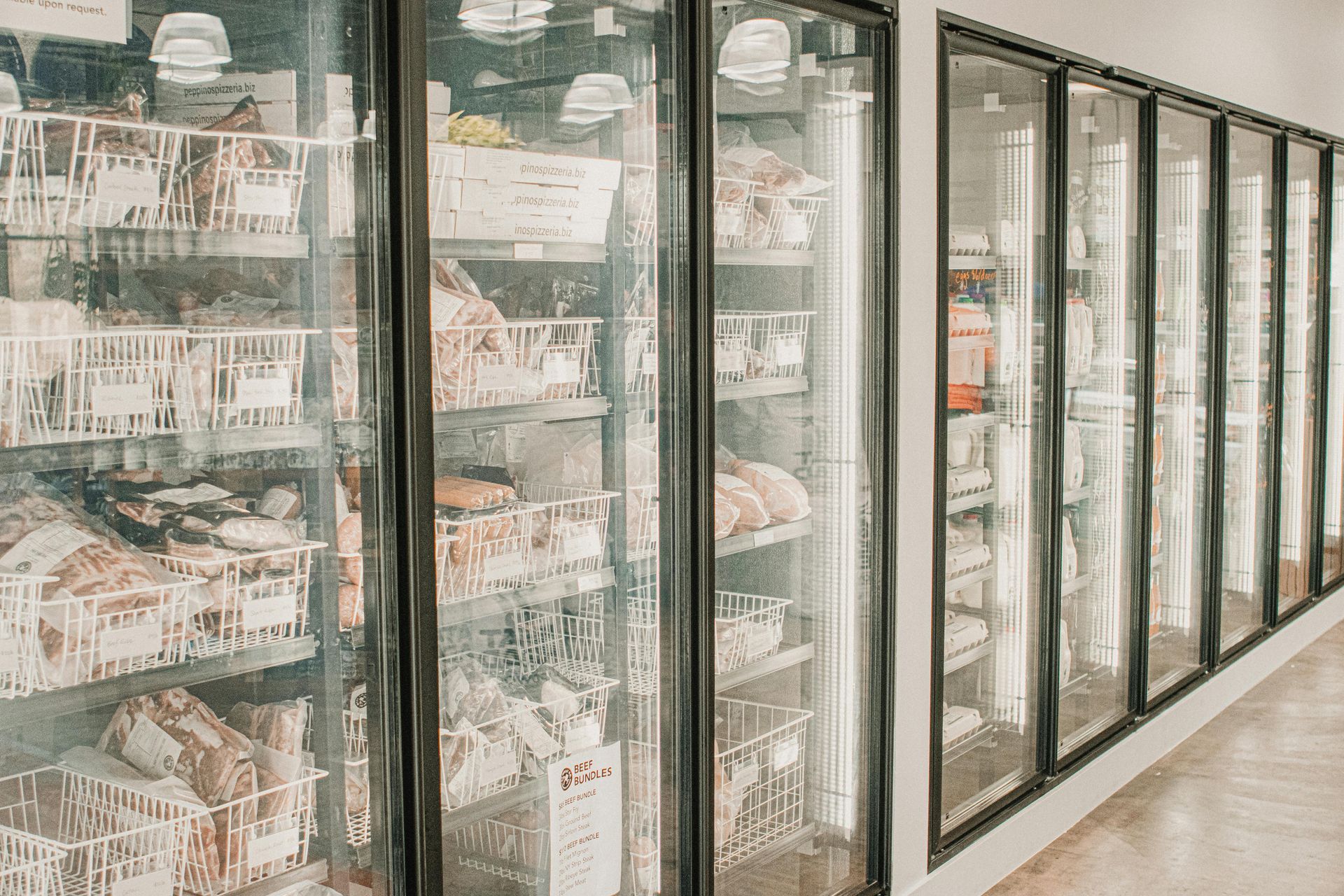 Freezer cases filled with meat products in a store. White baskets and clear doors line the wall.