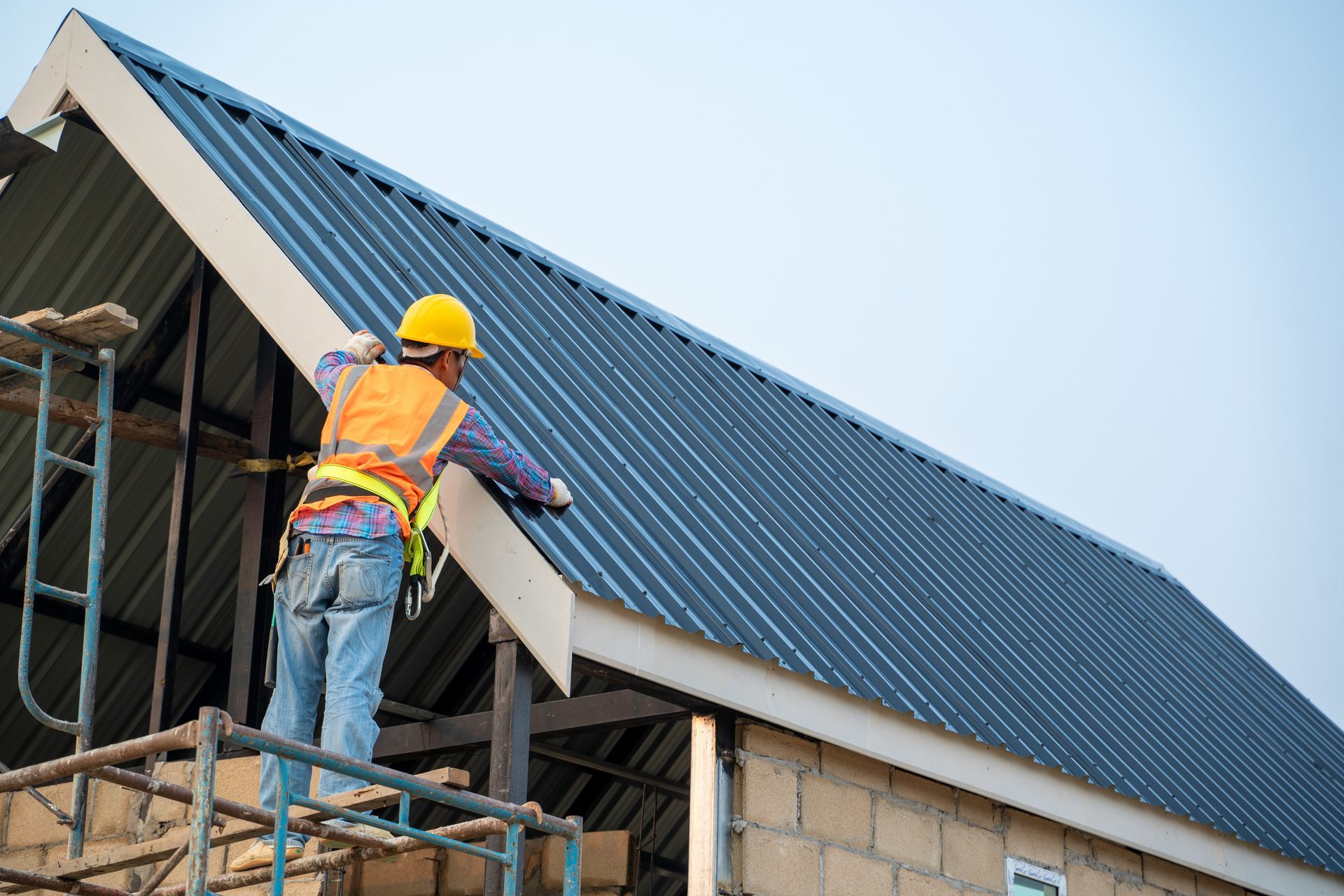 Roofer on scaffolding installing a dark corrugated metal roof, wearing safety gear, on a new construction with a light blue sky. Roofer on scaffolding installing a dark corrugated metal roof, wearing safety gear, on a new construction with a light blue sky.