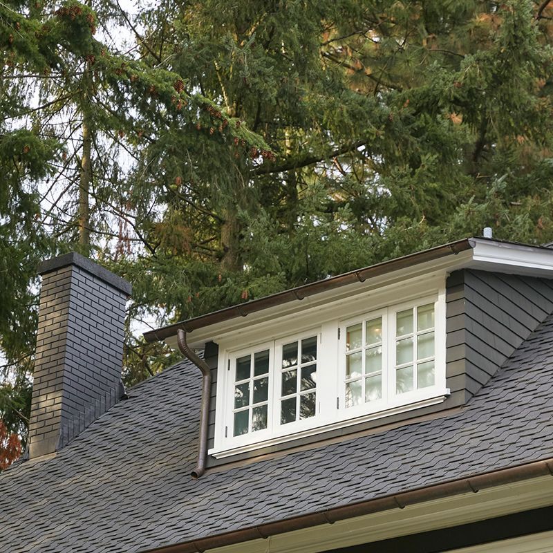 Dormer window on a dark shingled roof with a brick chimney, white trim, and green trees. Dormer window on a dark shingled roof with a brick chimney, white trim, and green trees.
