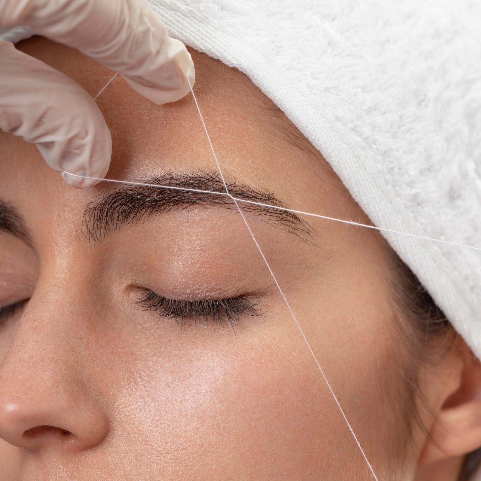 Gloved hand threading a facial thread along a closed eyebrow for cosmetic hair removal