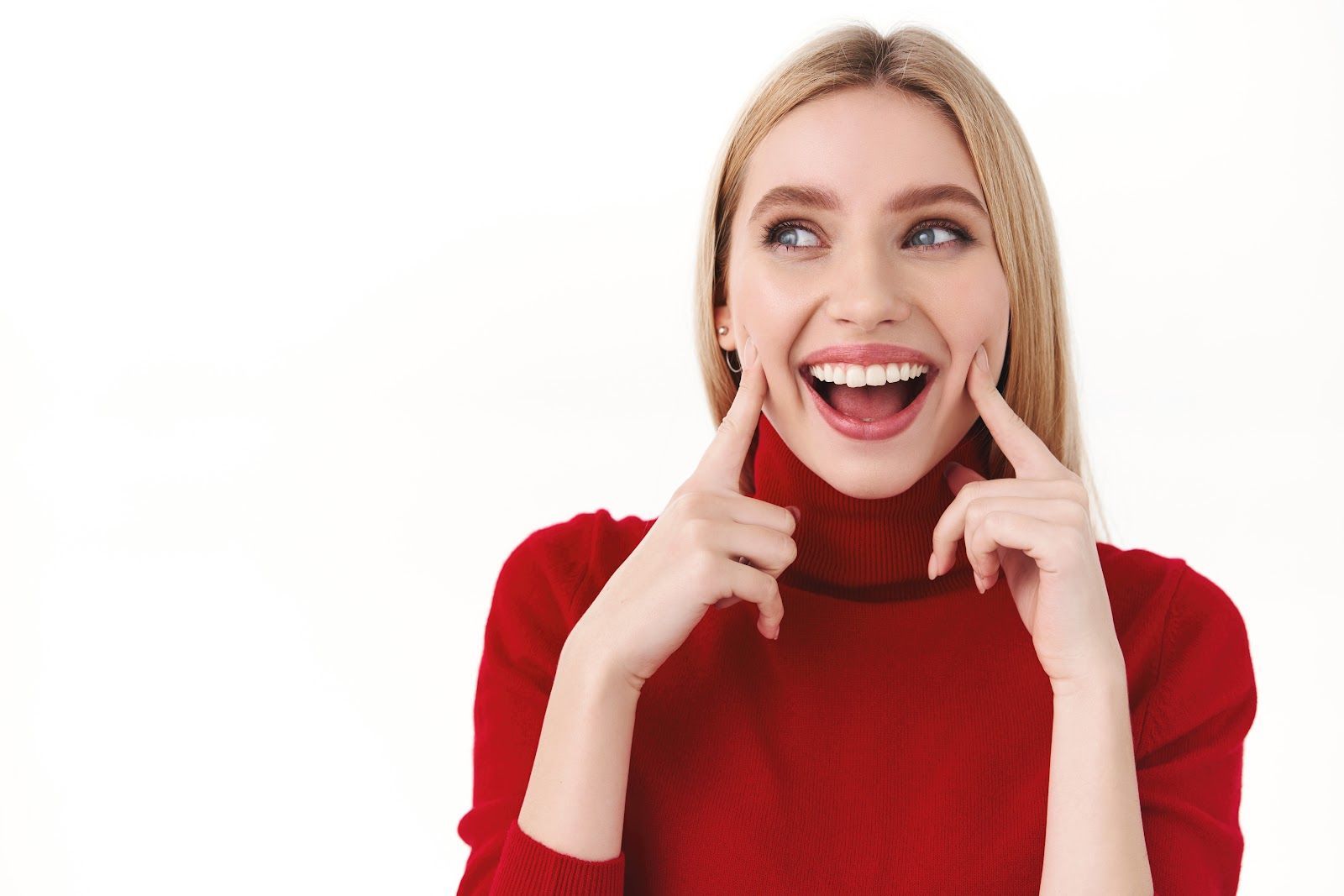 Smiling blonde woman in a red sweater pointing at her cheeks against a white background