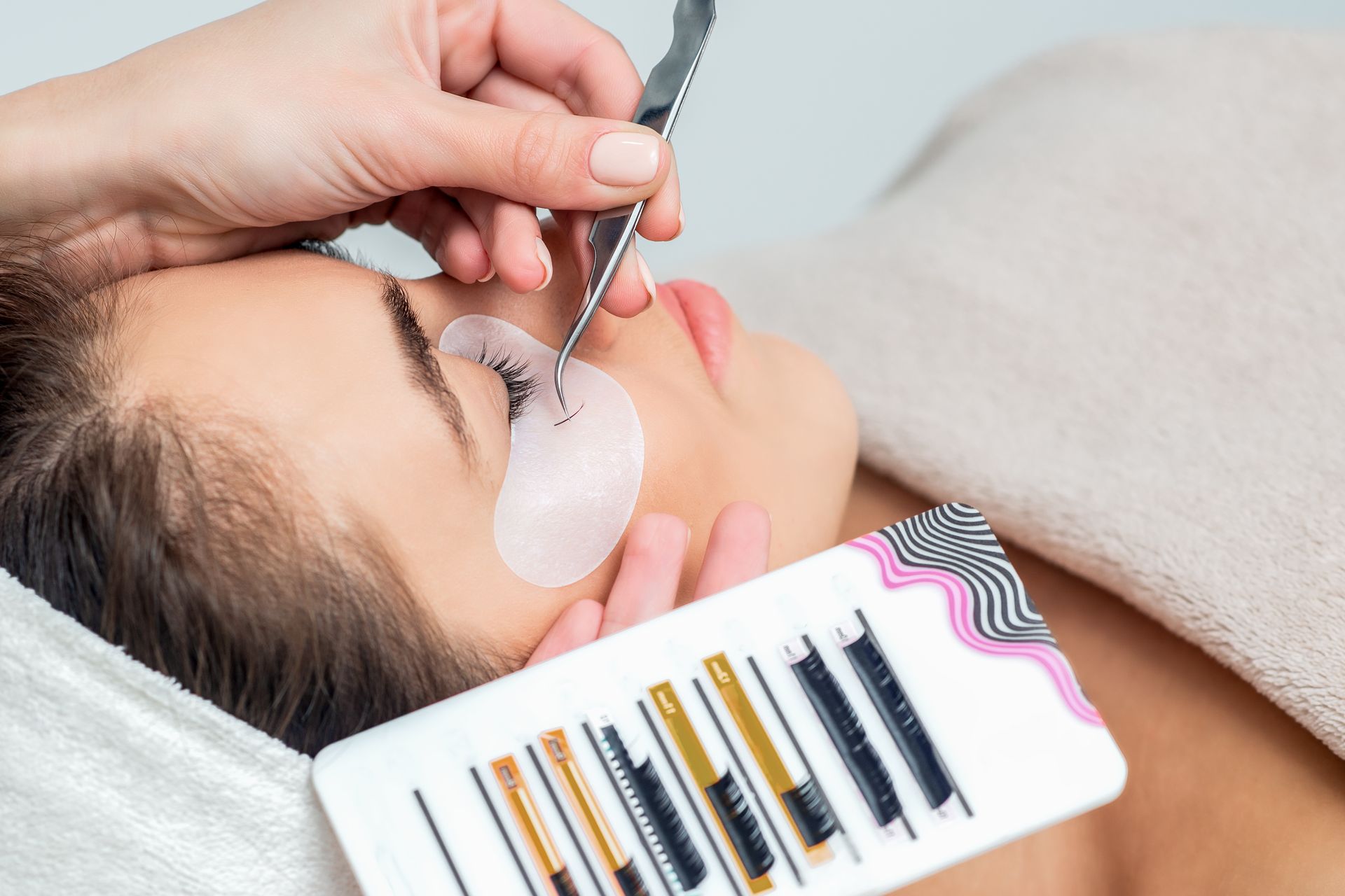 Eyelash extension treatment with technician using tweezers on a client lying down, lash tray nearby