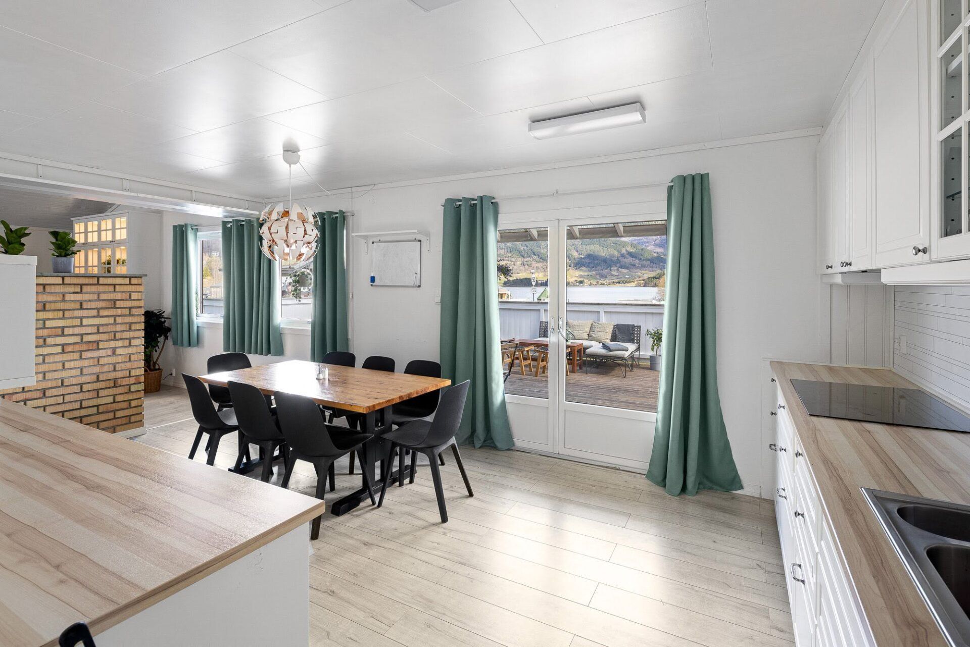 Bright dining room and kitchen with a wooden table, black chairs, teal curtains, and a view of the water