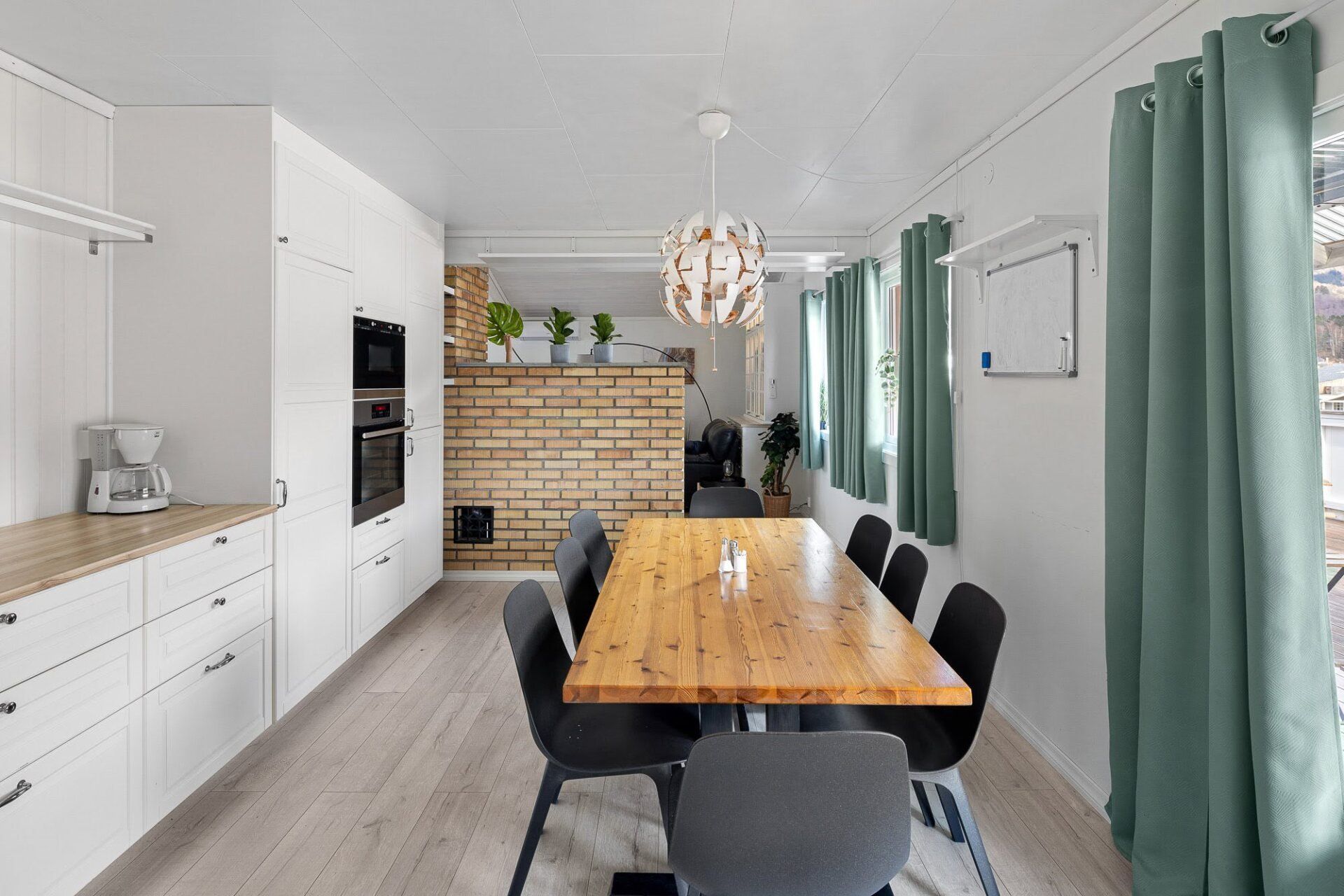 Bright modern dining room with wooden table, black chairs, white cabinets, green curtains, and brick fireplace