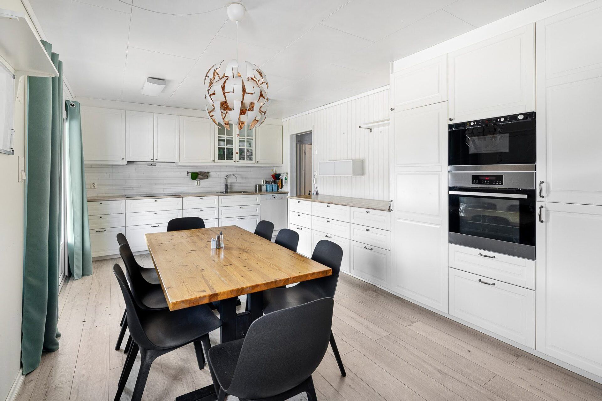 Bright modern kitchen-dining room with wooden table, black chairs, white cabinets, and built-in ovens.