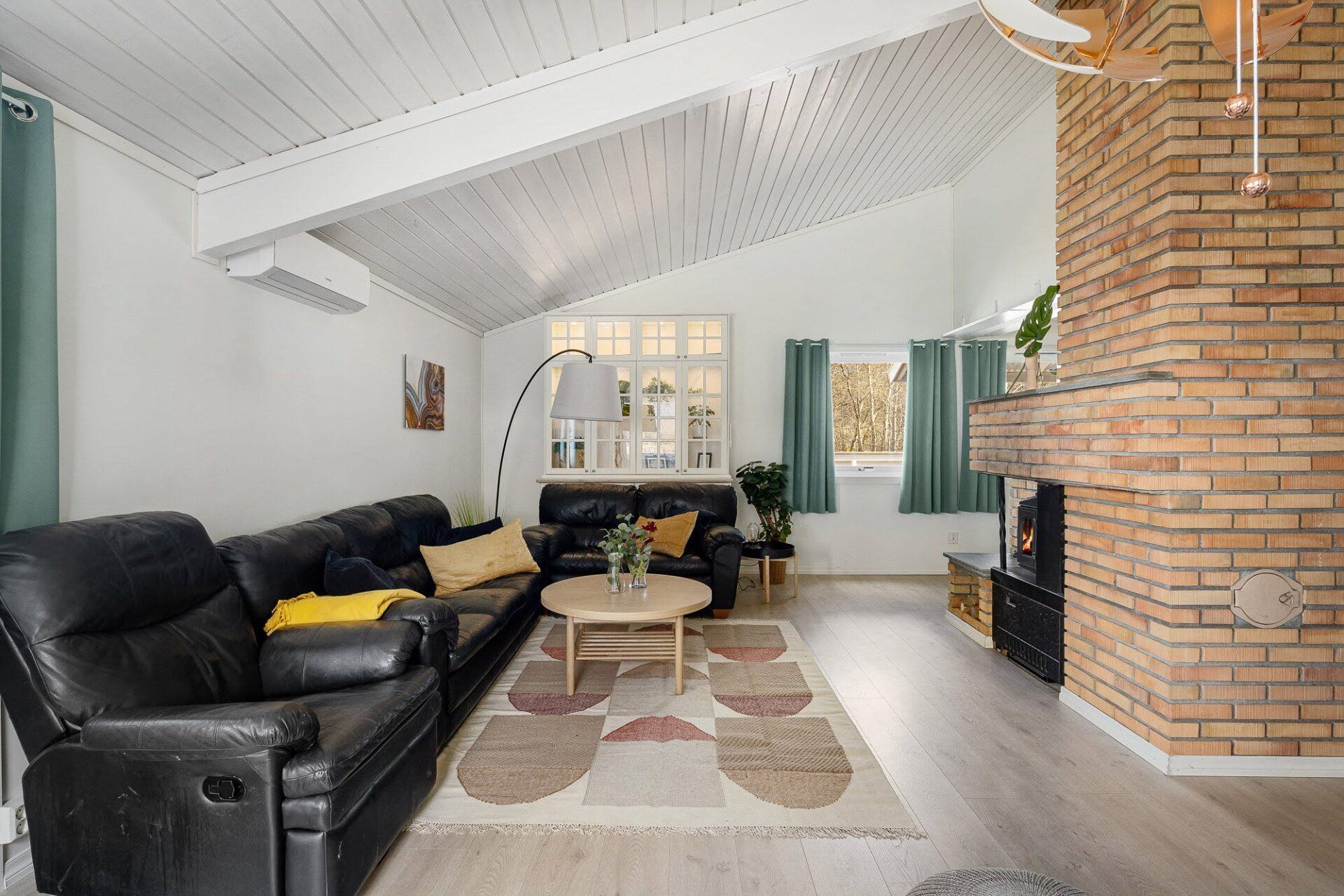 Bright living room with black sofas, patterned rug, and a brick fireplace under a vaulted white ceiling