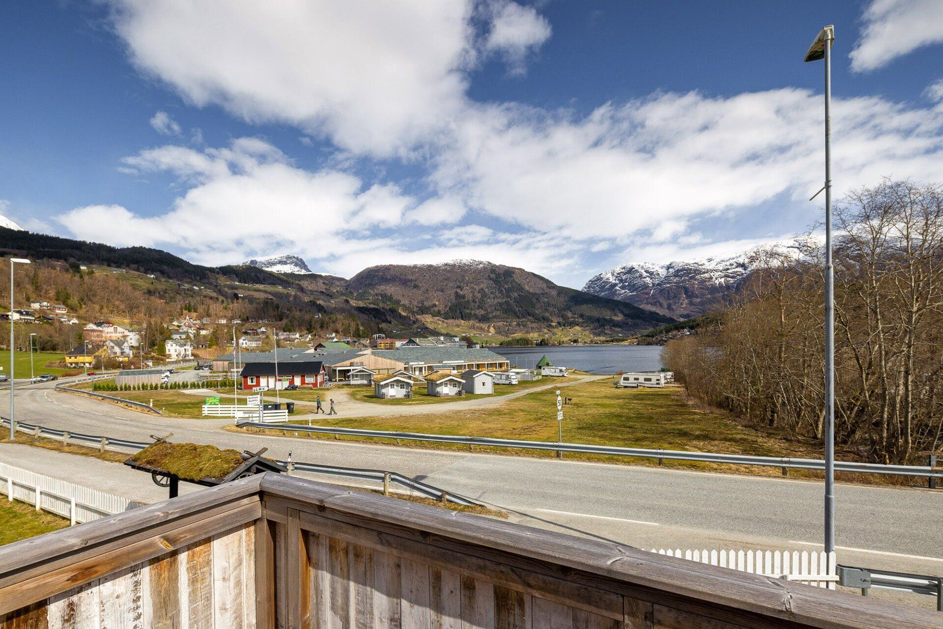 Wooden deck overlooking a small village, road, lake, and snow-capped mountains under a cloudy sky