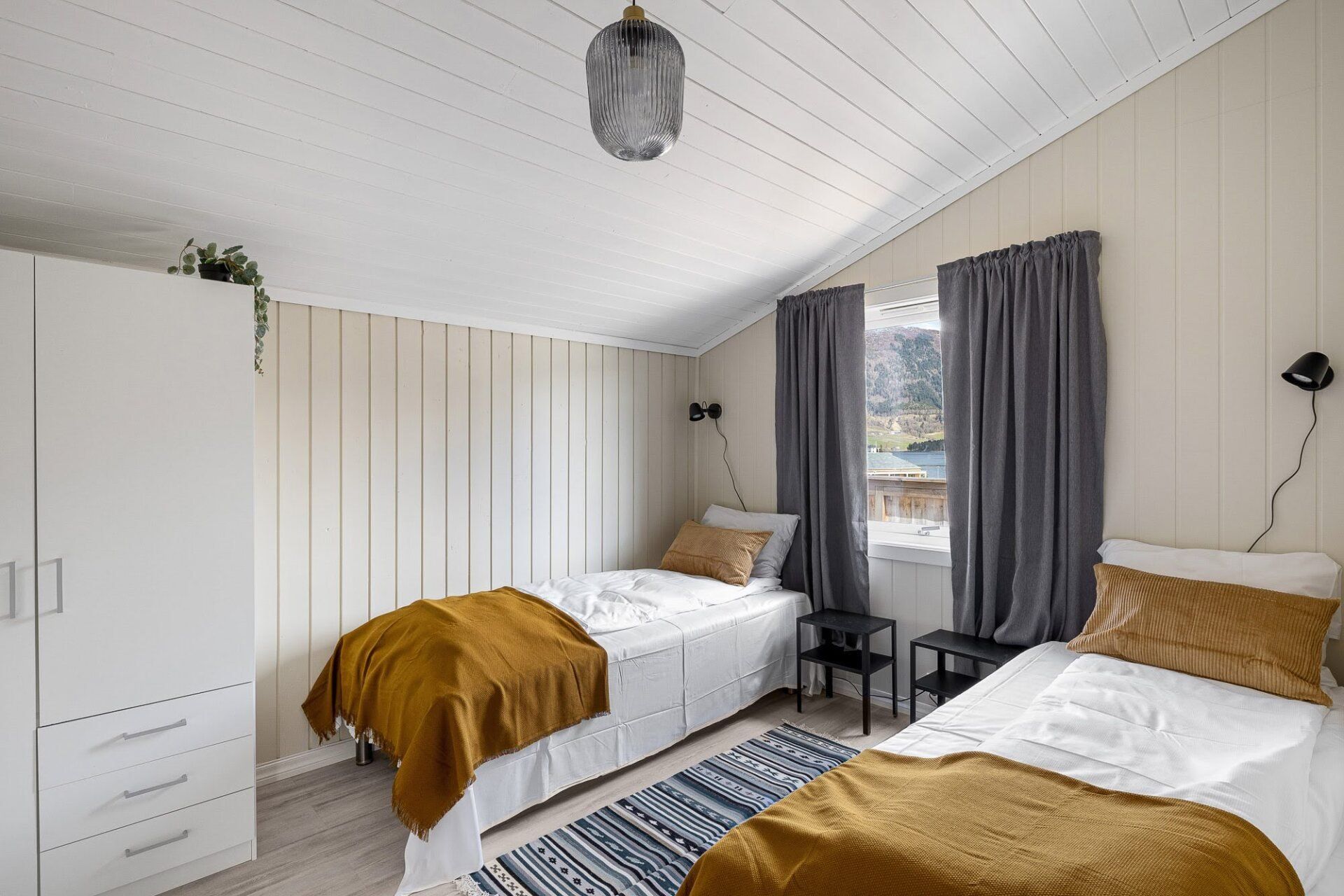 Bright bedroom with two single beds, white wardrobe, gray curtains, and striped rug