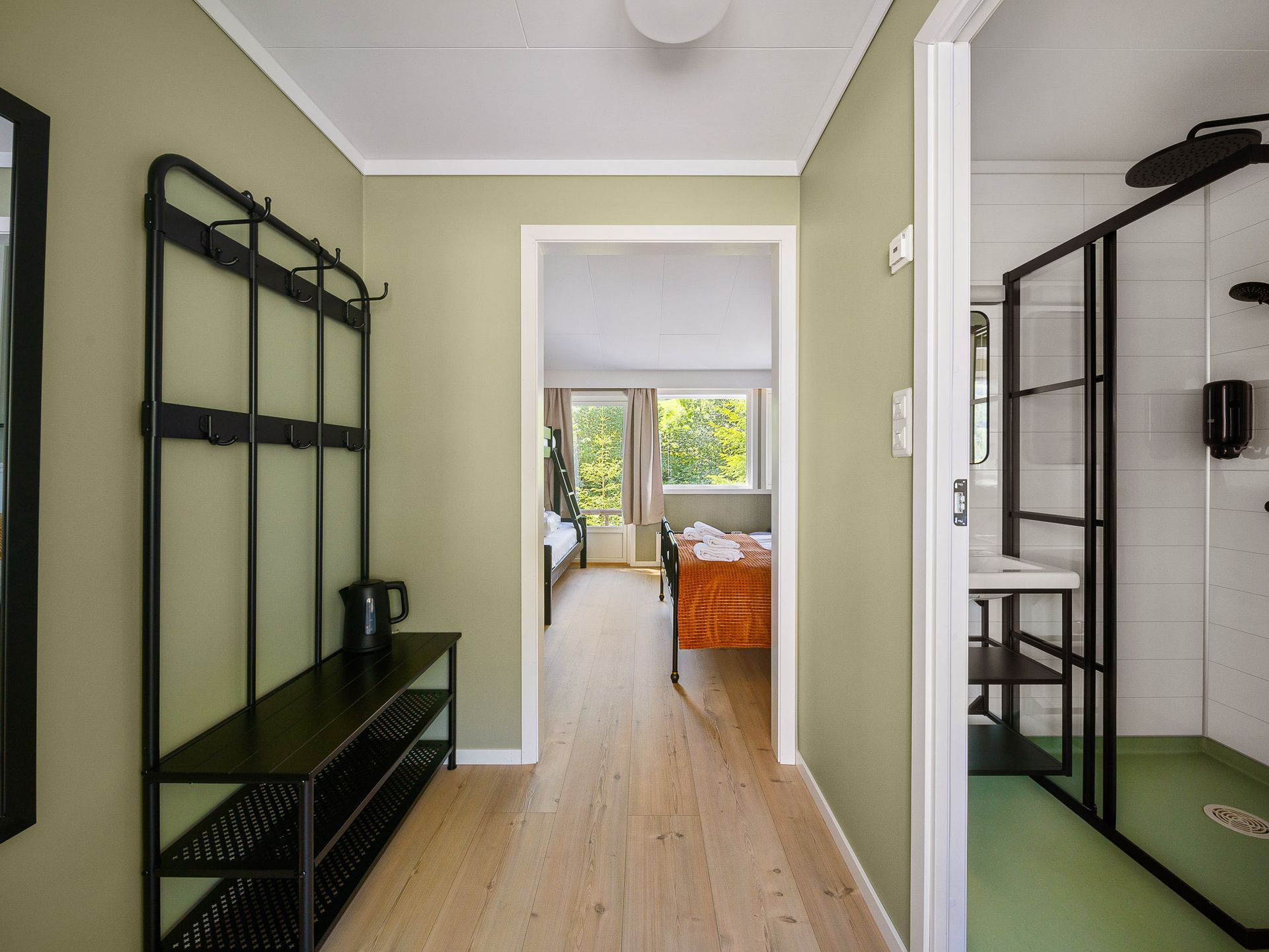 Narrow hallway with black shelving, pale walls, wood floor, and a view into a bright room with orange chairs.