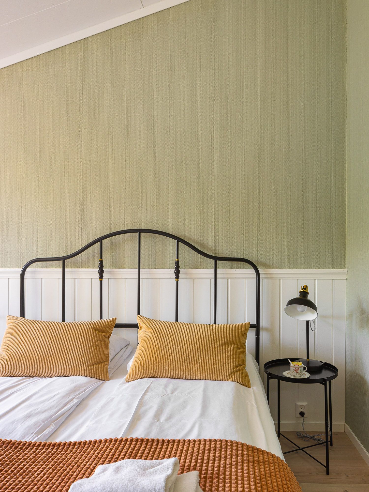 Cozy bedroom with black iron bed, white and mustard bedding, and green patterned wall above white wainscoting