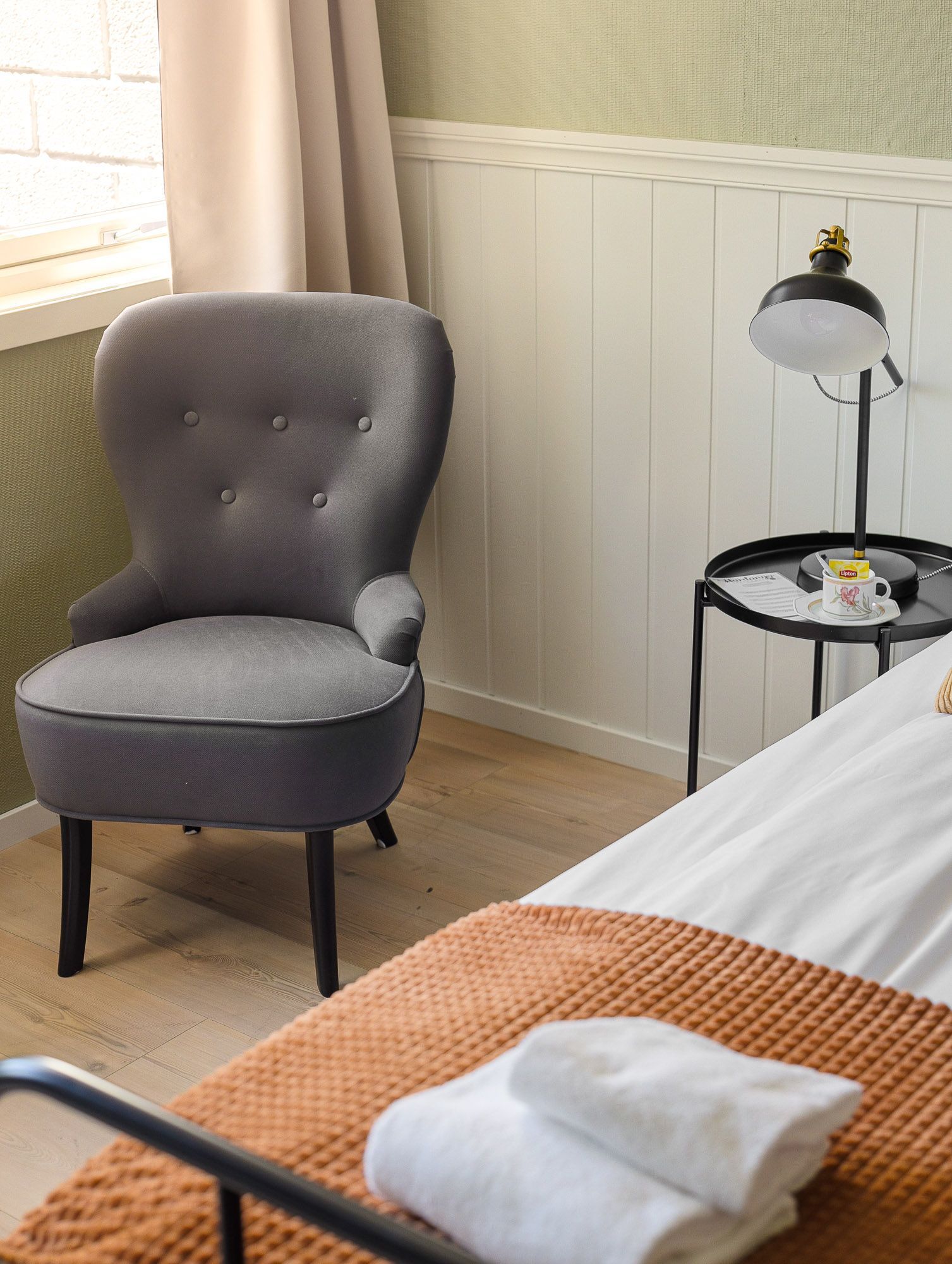 Cozy bedroom corner with gray tufted chair, bedside lamp, and folded towels on a bed in the foreground