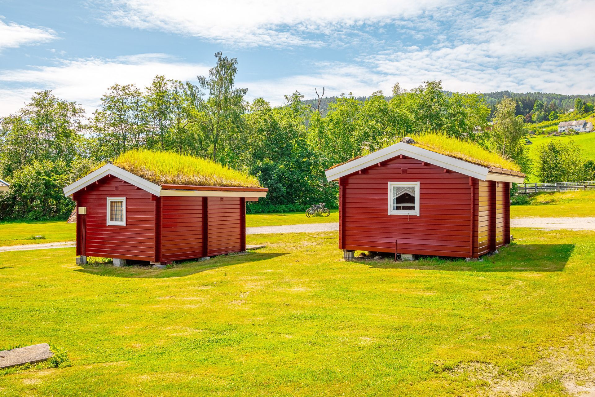 Two red wooden cabins on a sunny green lawn beside a lake and trees