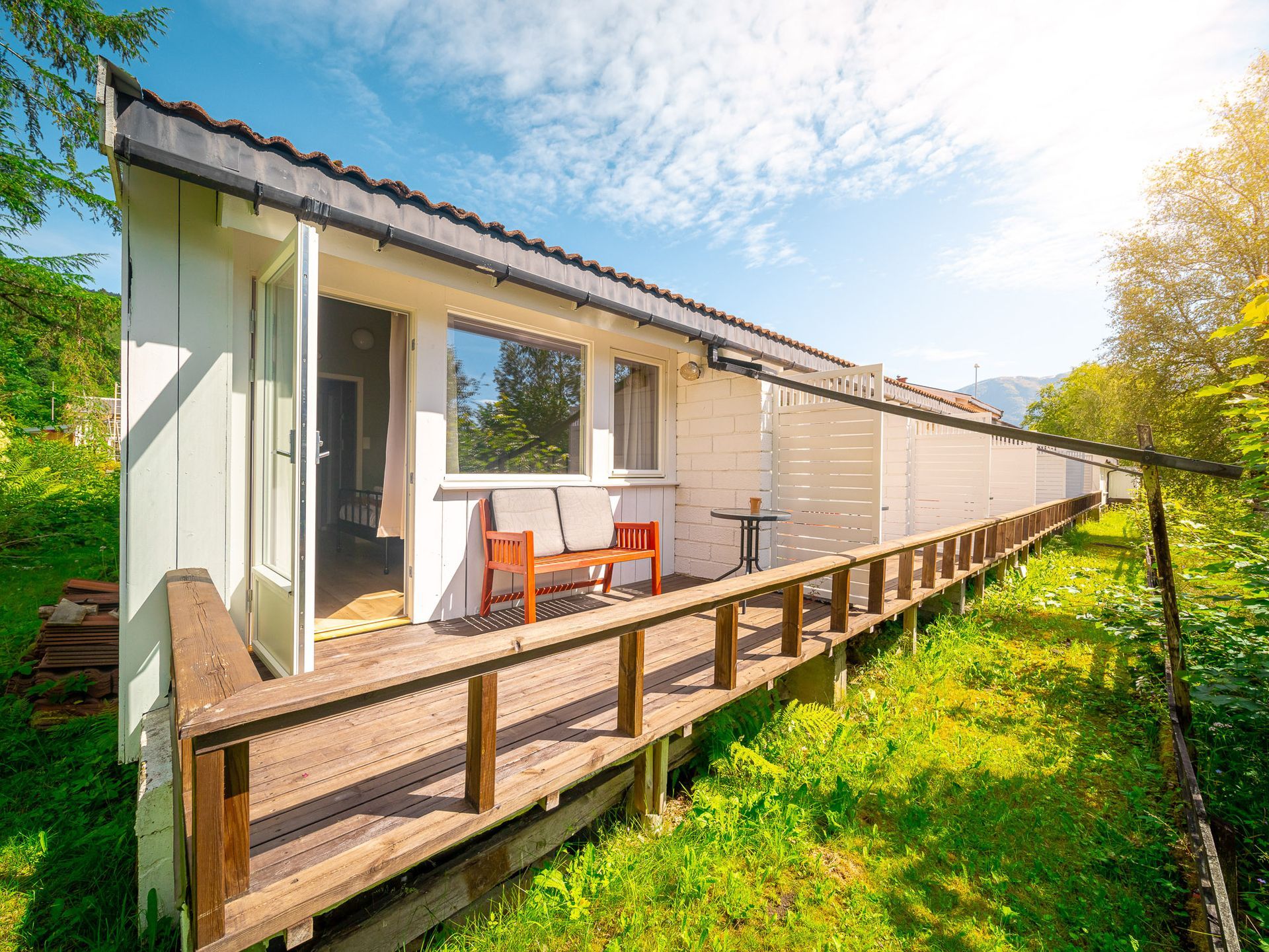 Single-story house with a wooden ramp deck and red chairs in a sunny garden.