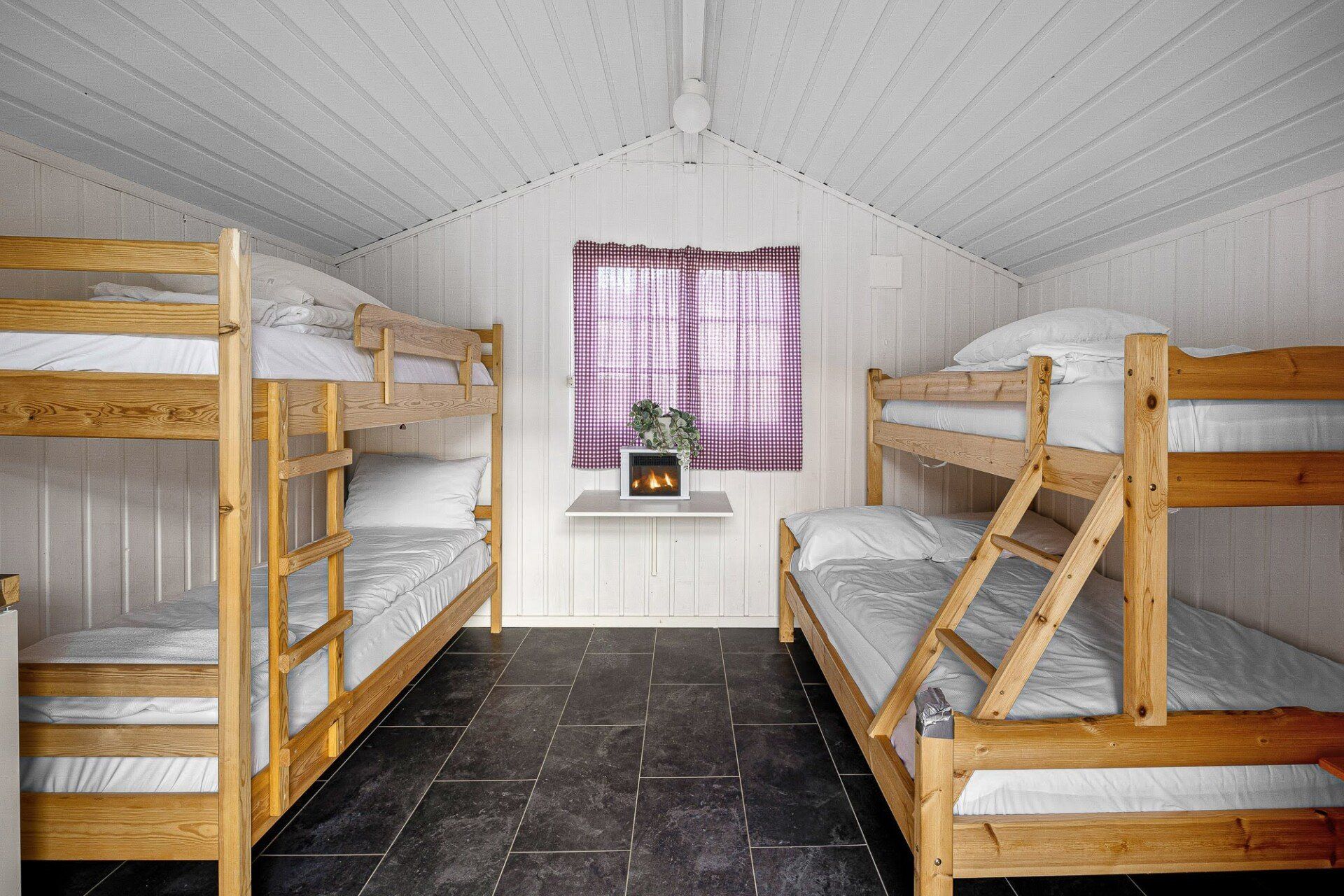 Small attic dorm room with two sets of bunk beds, white walls, dark floor, and a window with red checkered curtains