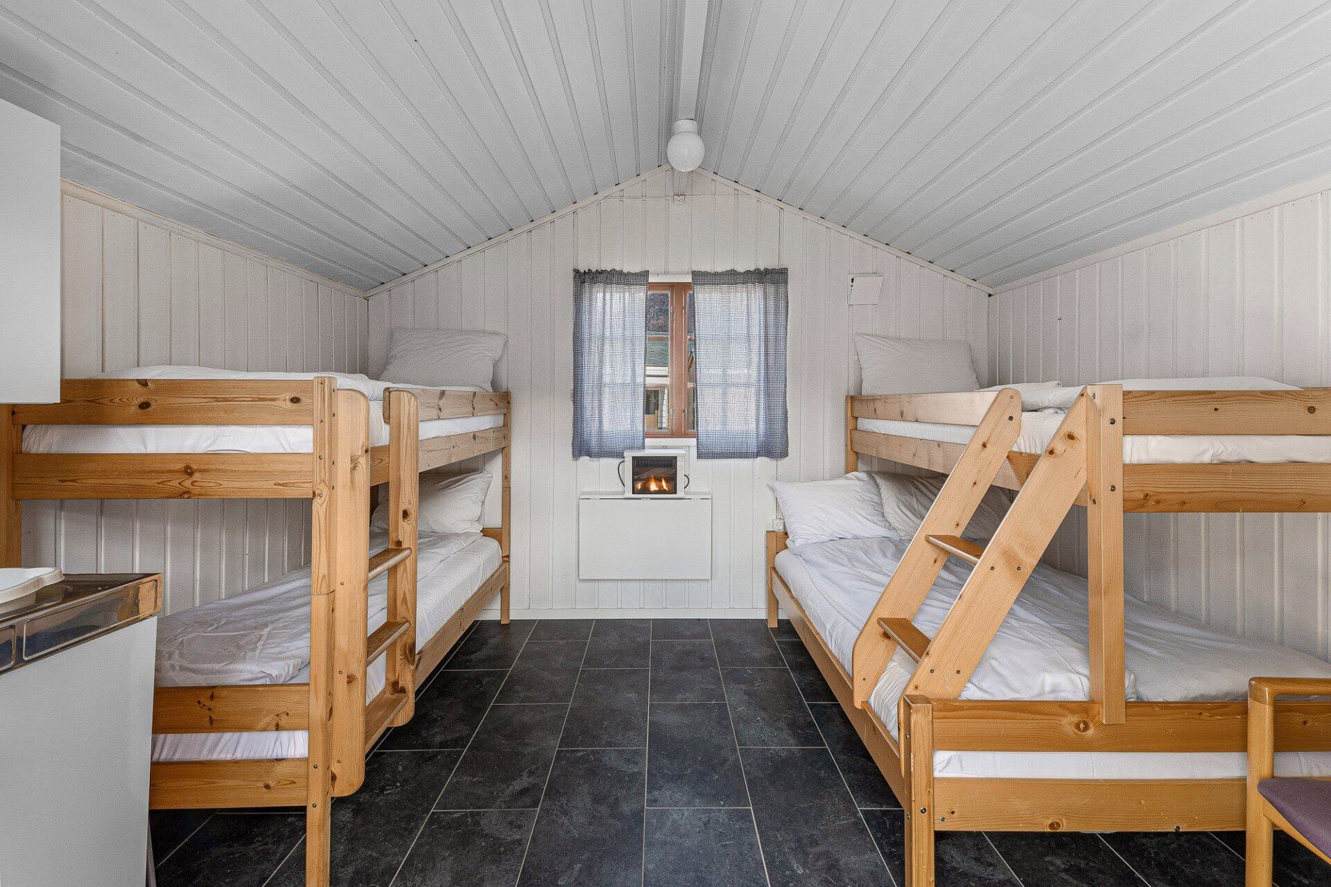 Small dorm-style bunk room with four wooden bunk beds, white walls, and a window at the far end
