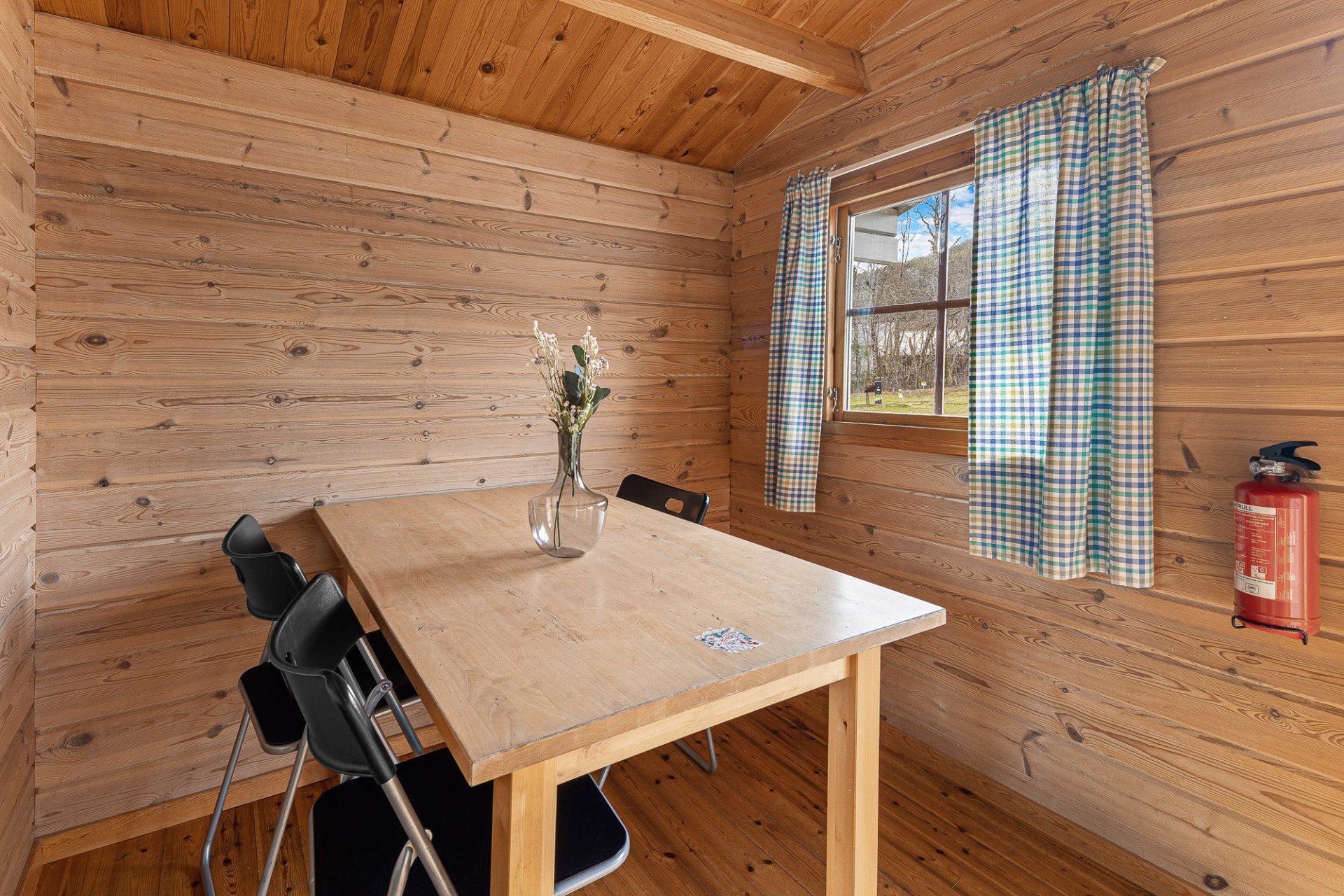 Small wooden cabin dining area with a table, chairs, window, and red fire extinguisher.